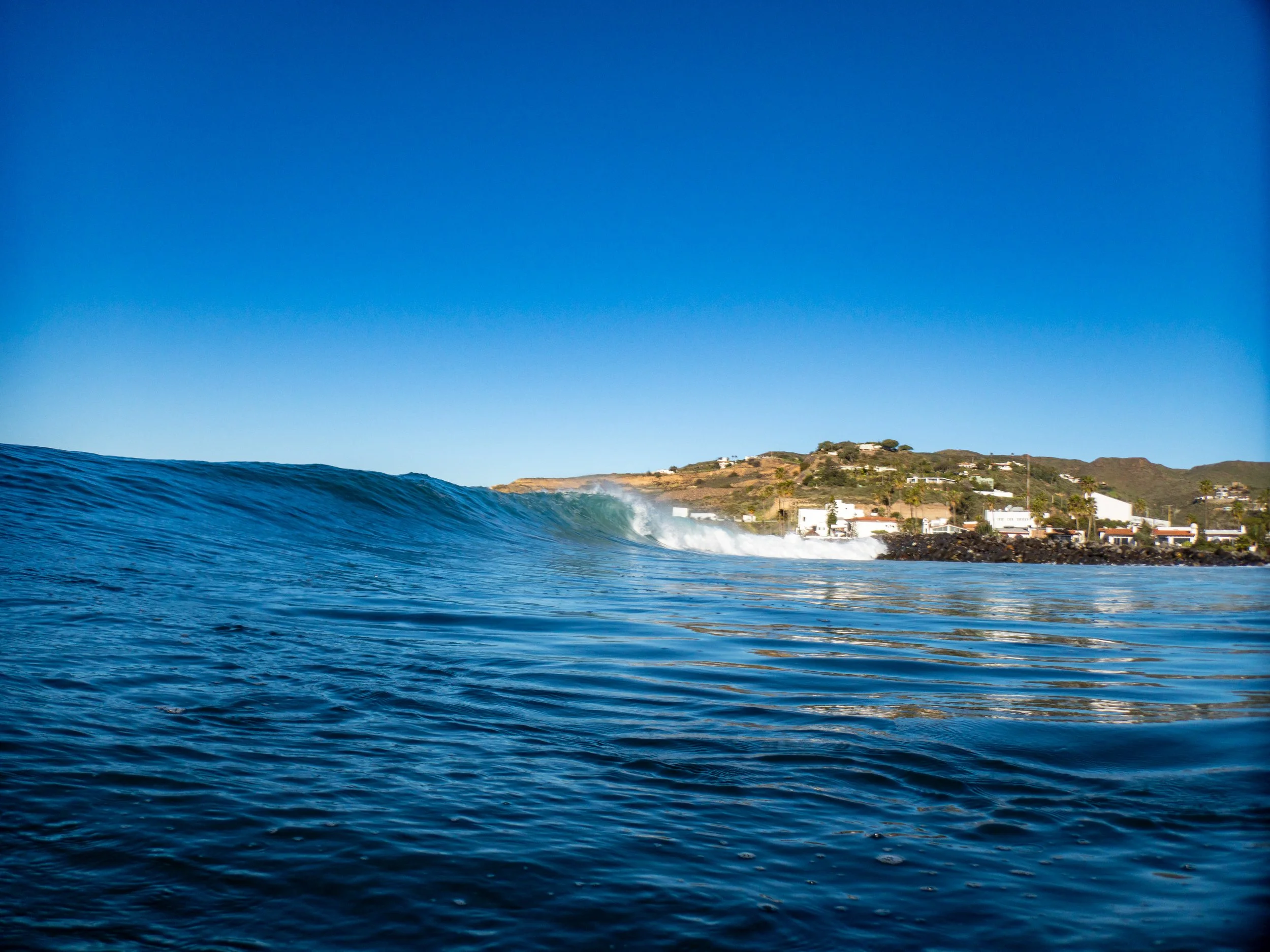 Ocean wave with waves crashing near a coastline with residential buildings and hills in the background under a clear blue sky.