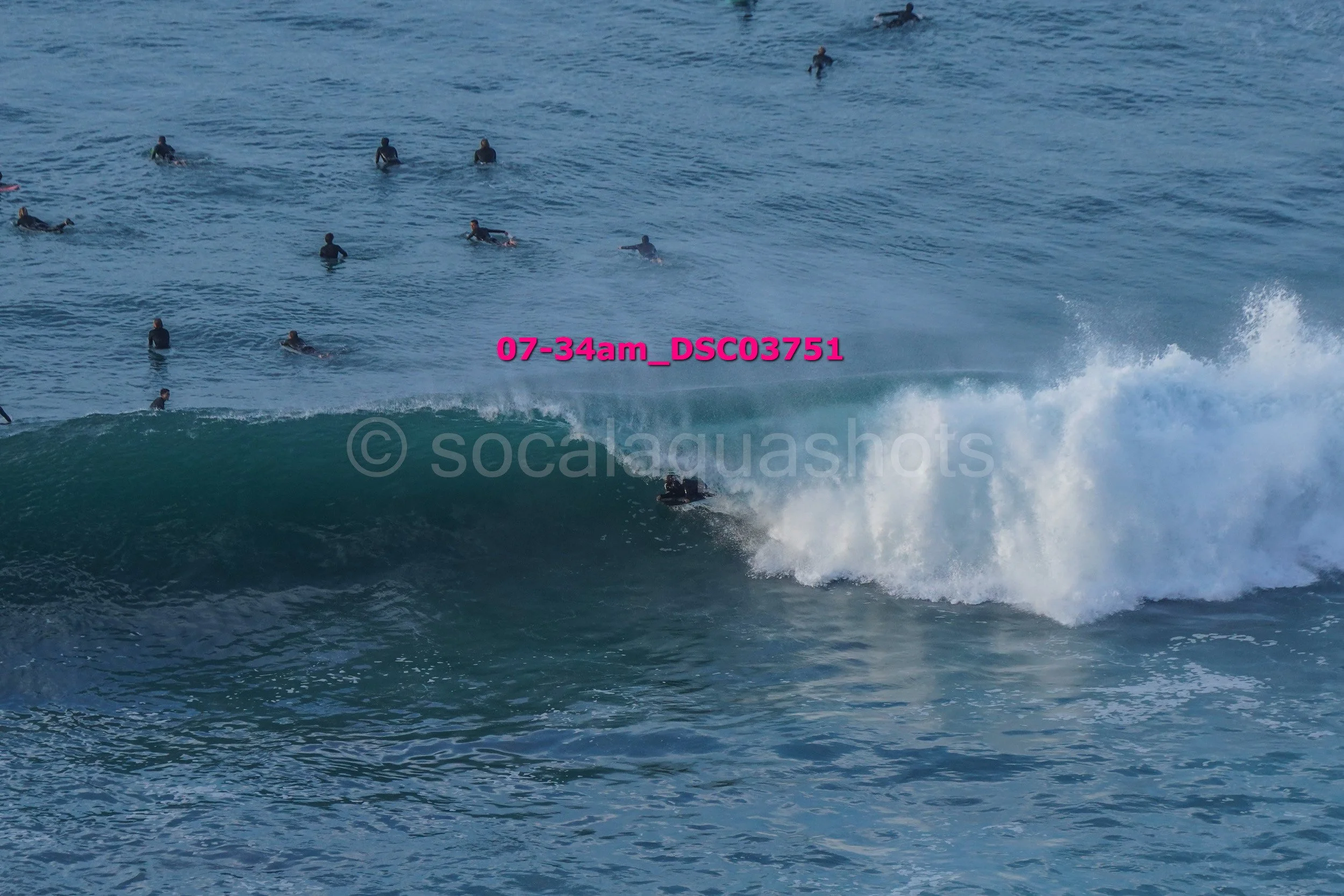 Surfers riding a wave in the ocean with many people in the water, some waiting and others swimming. The image shows the beach scene with a focus on the surfer on the wave.