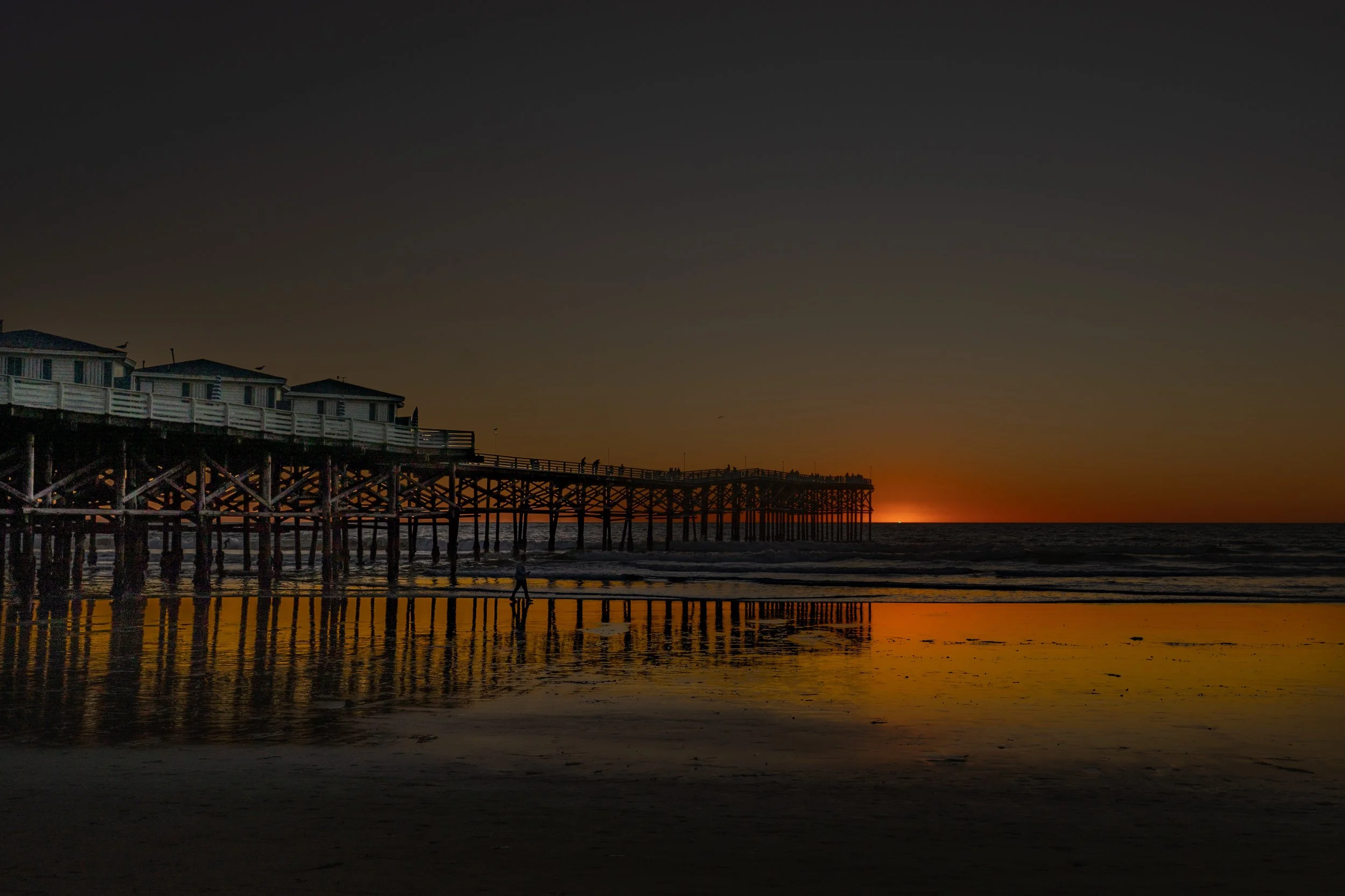 Sunset over a pier extending into the ocean with reflections on wet sand.