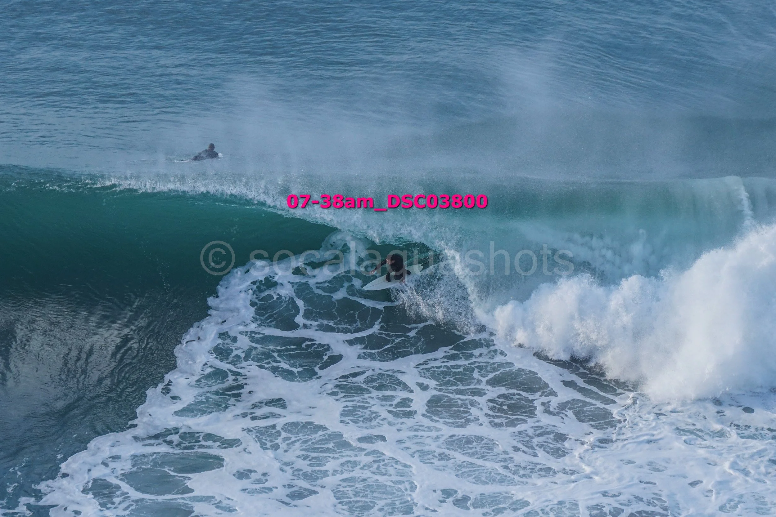 A surfer riding a large ocean wave with a second surfer in the background.