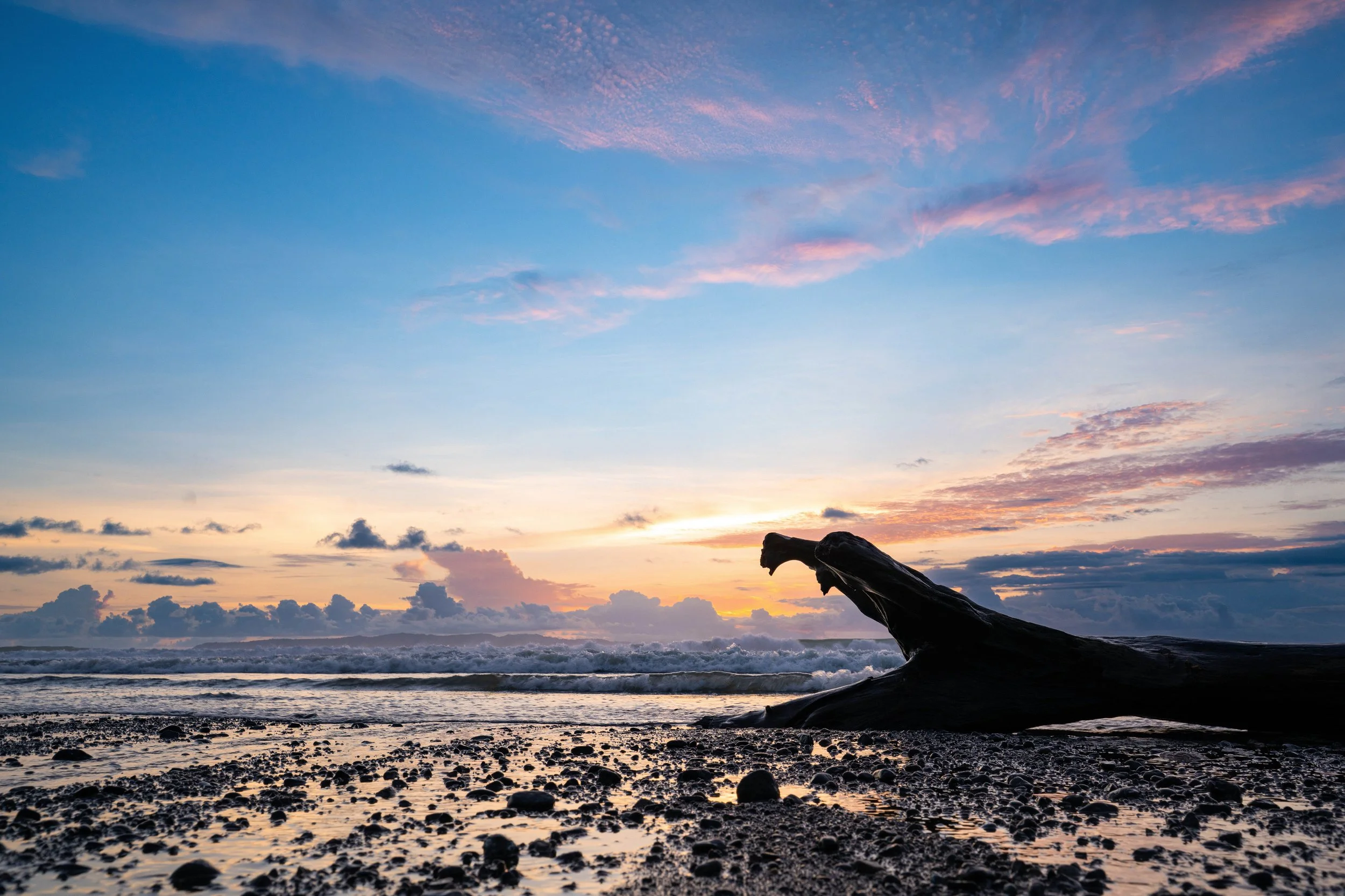 A sunset over the ocean with a rocky beach, clouds, and a piece of driftwood.