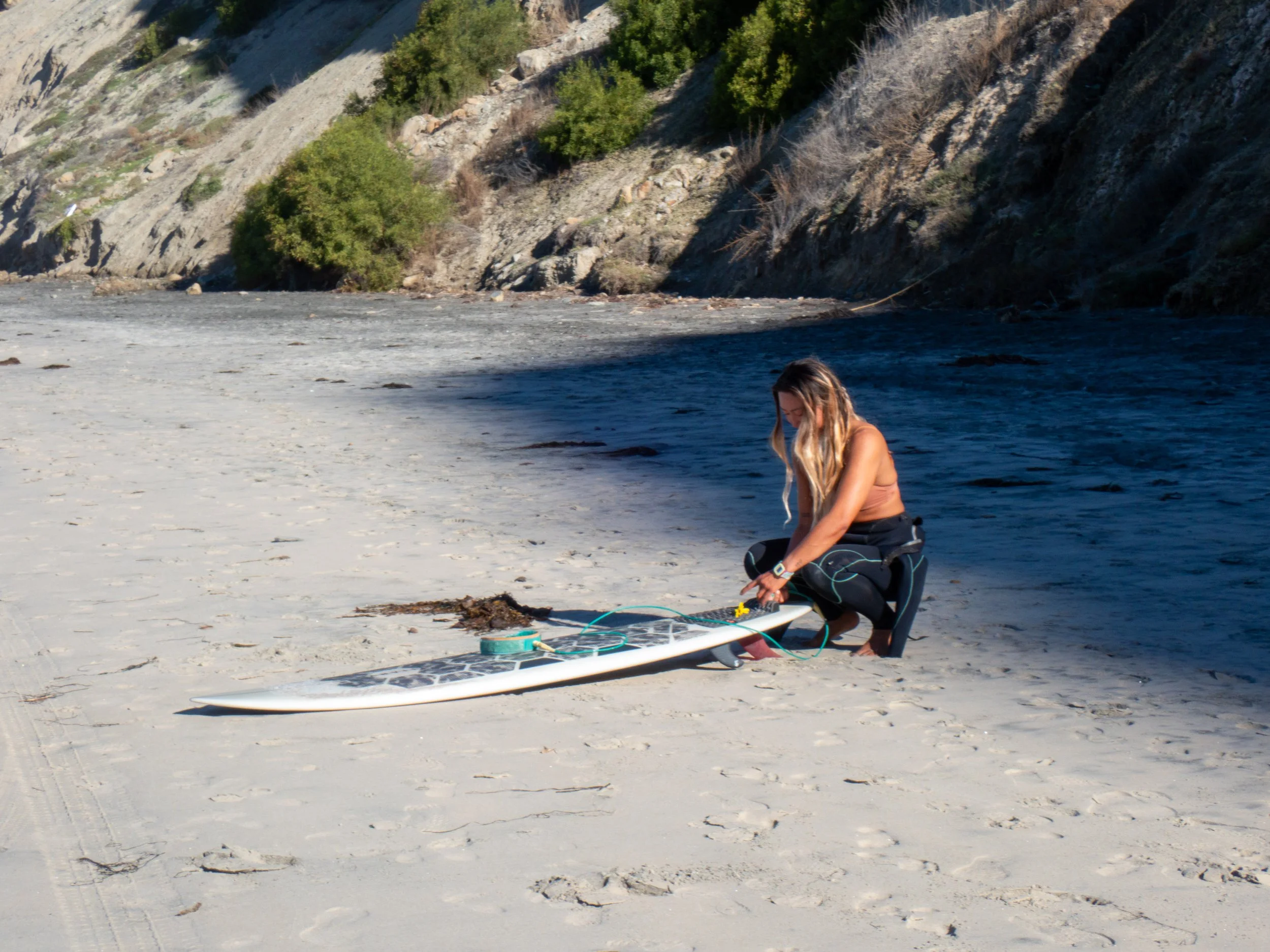 A woman kneeling on a sandy beach next to a river, preparing her stand-up paddleboard.