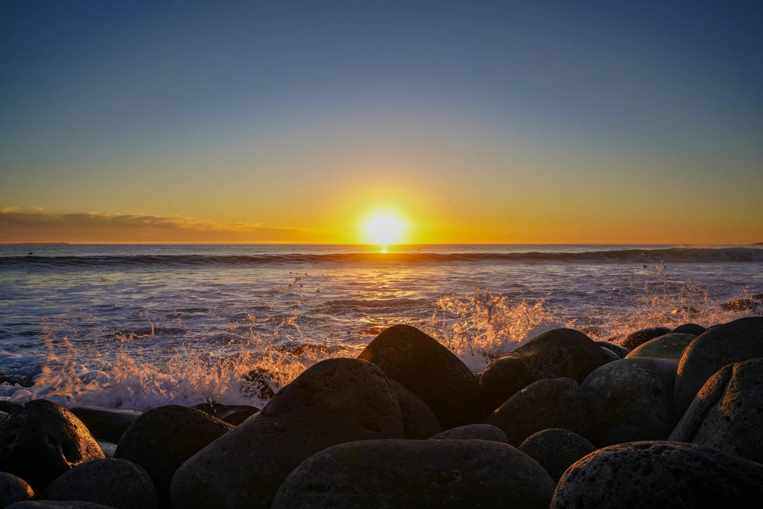 A sunset over the ocean with rocks in the foreground and waves crashing against them.