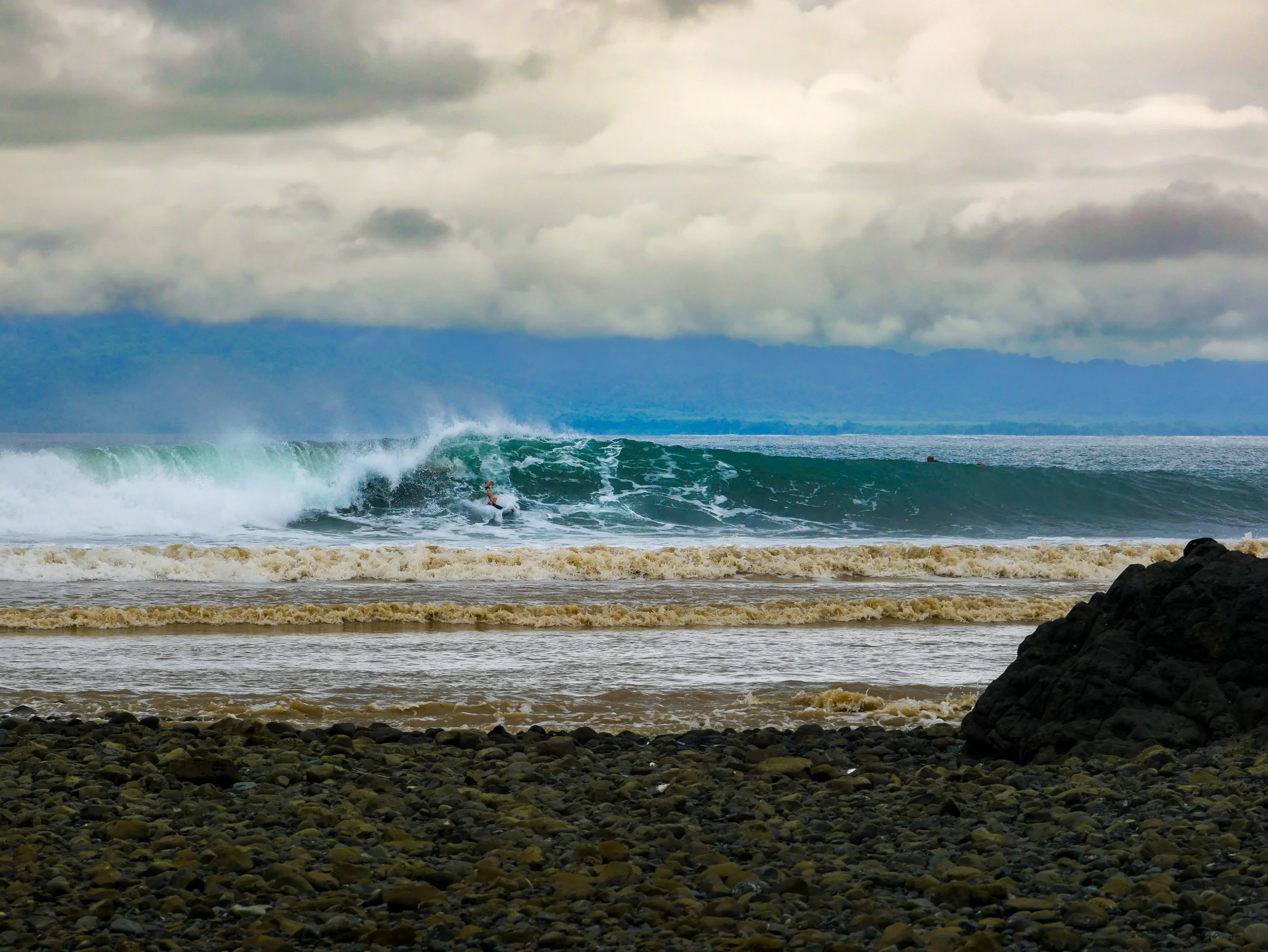 Surfer riding a wave near rocky beach with stormy sky