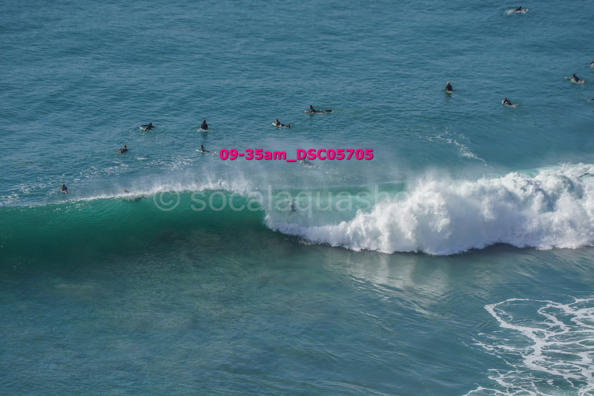 Multiple surfers in wetsuits sitting on surfboards in the water, waiting for waves, with a large breaking wave in the foreground.