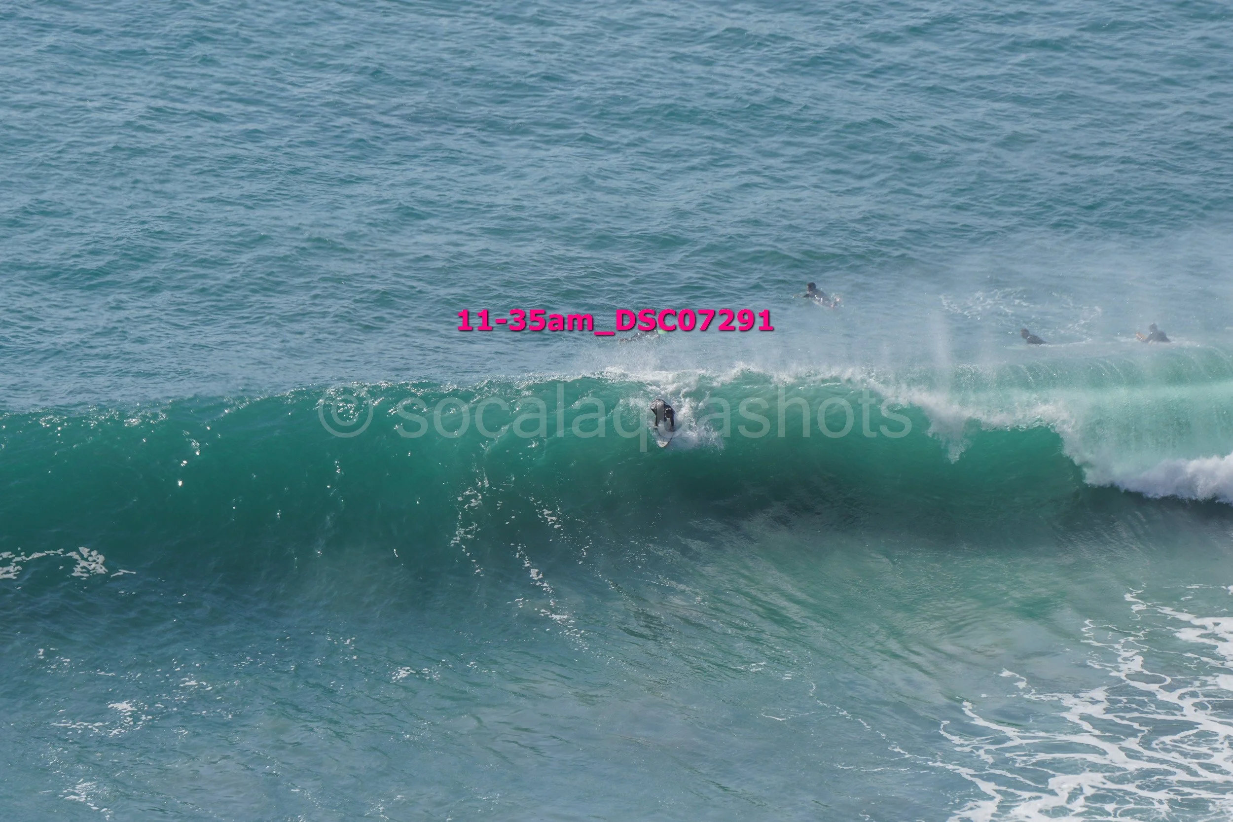A surfer riding a large wave with several other surfers visible in the background in the ocean.
