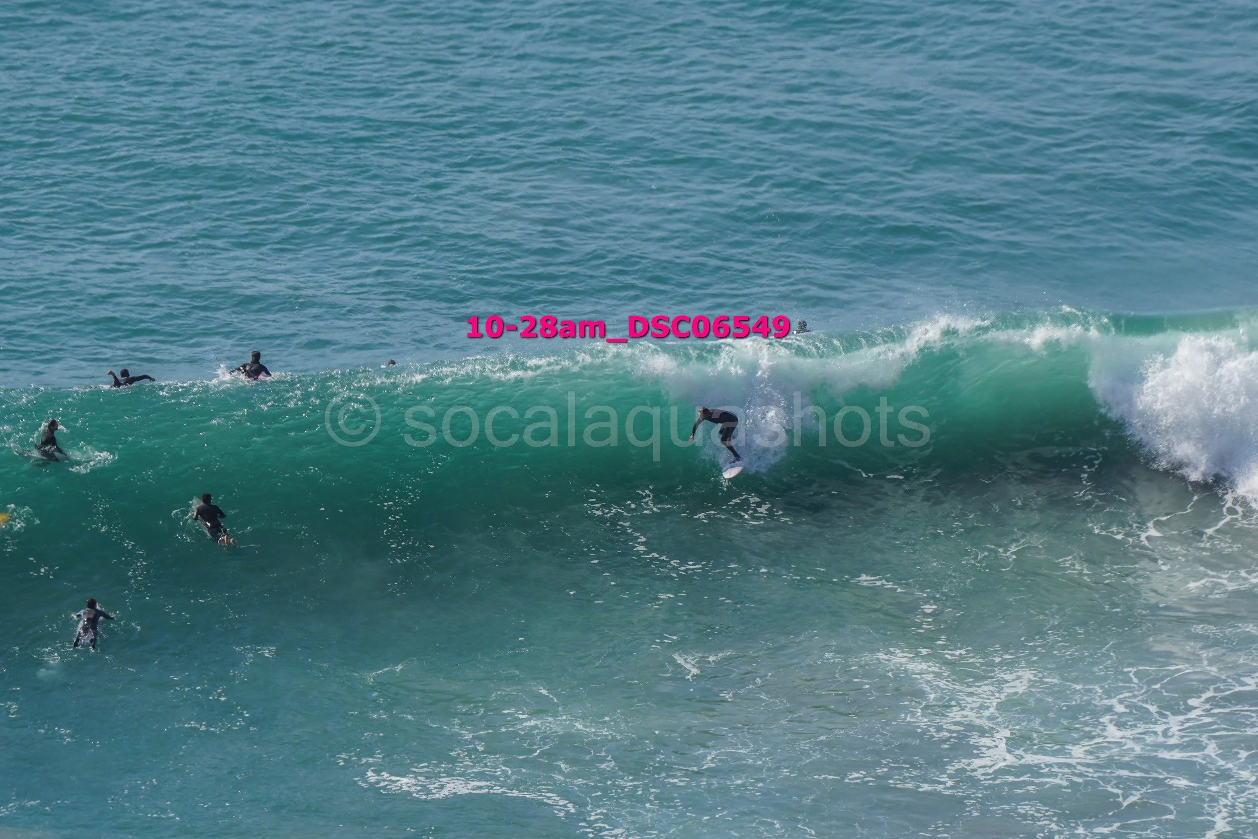 A person surfing a large ocean wave with several others swimming nearby in the water.