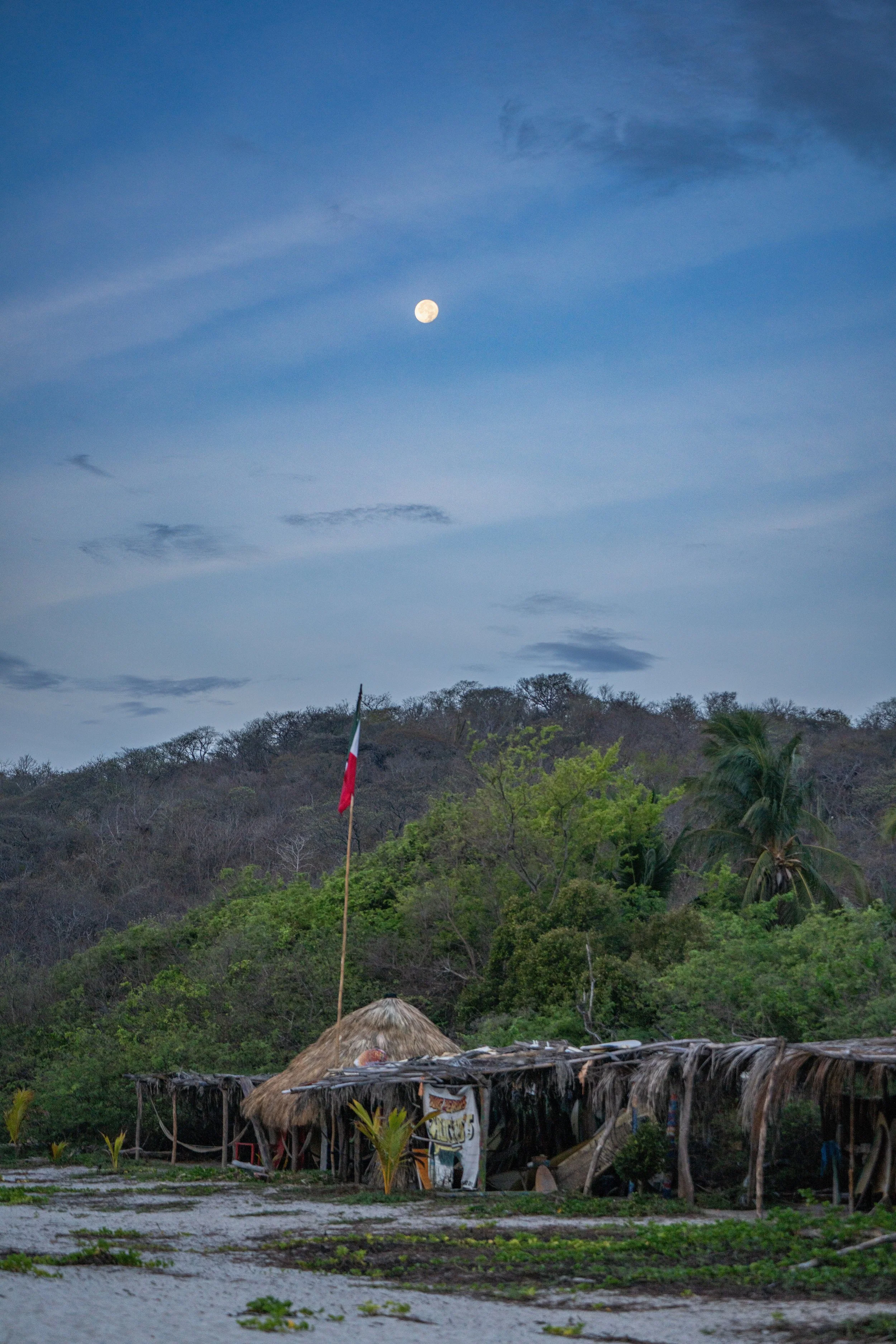 A rural scene with a thatched roof structure in the foreground, lush green trees on rolling hills in the background, and the moon visible in a cloudy sky.