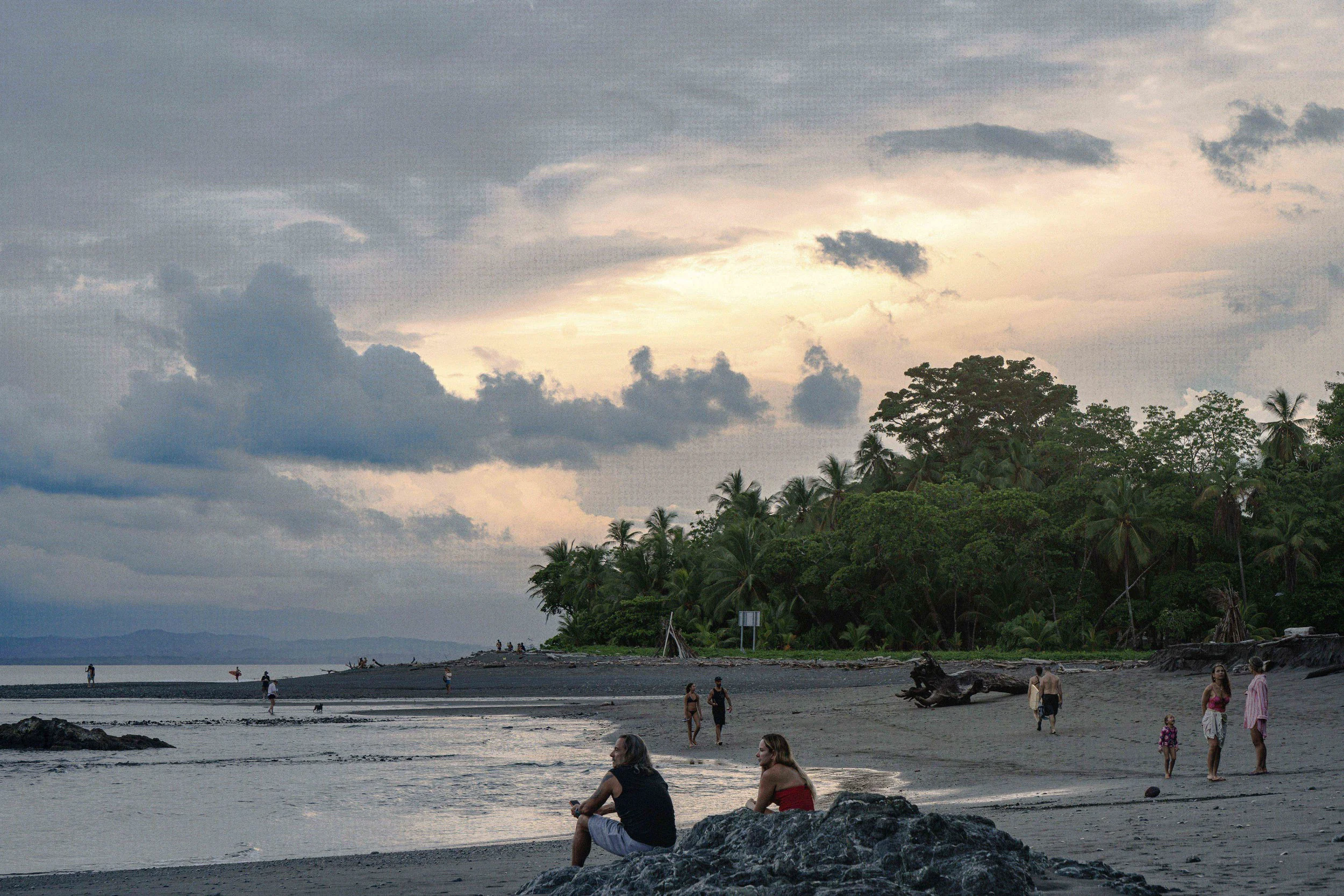 People strolling and sitting on a beach shoreline with palm trees and lush greenery, cloudy sky overhead at sunset.