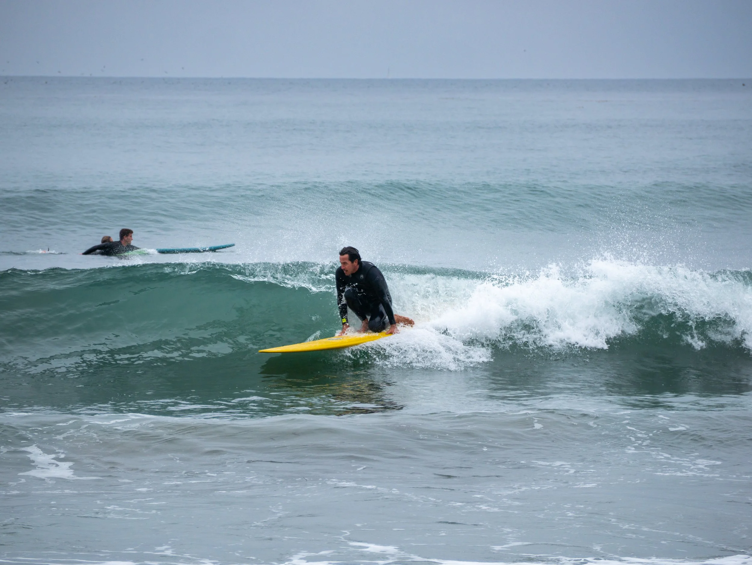 A man in black wetsuit surfing on a yellow surfboard on a small wave, with two other surfers in the background on the ocean.