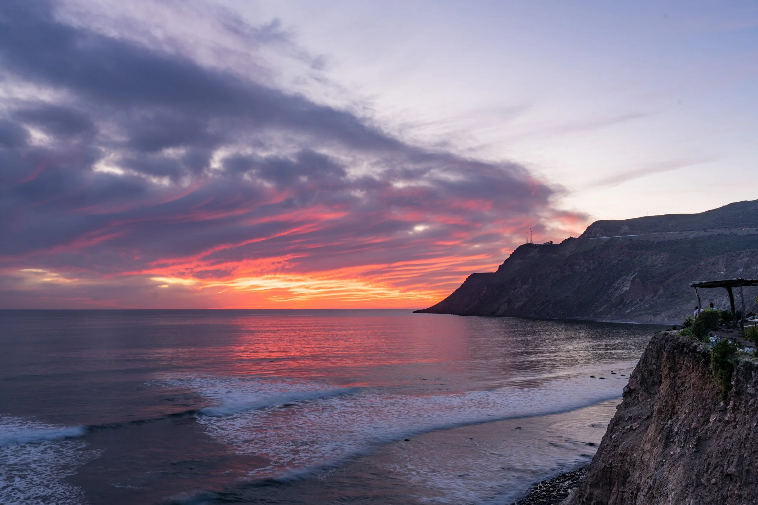 Sunset over a calm ocean near a rugged coastline with cliffs and a small structure on the right.