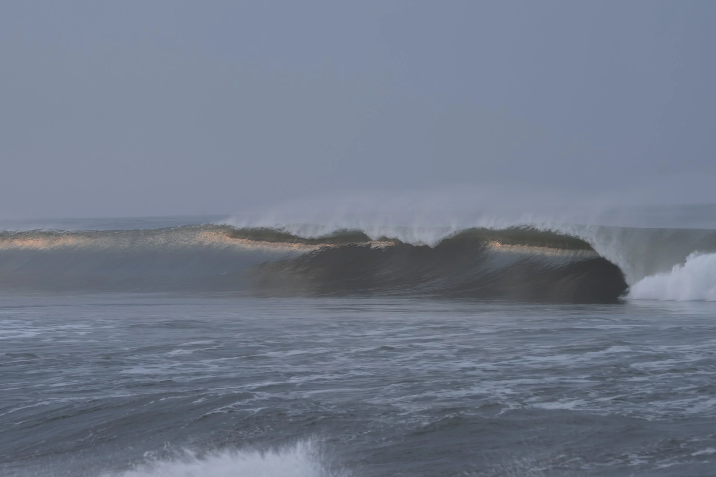 Ocean waves crashing with a large wave in the distance under a cloudy sky.