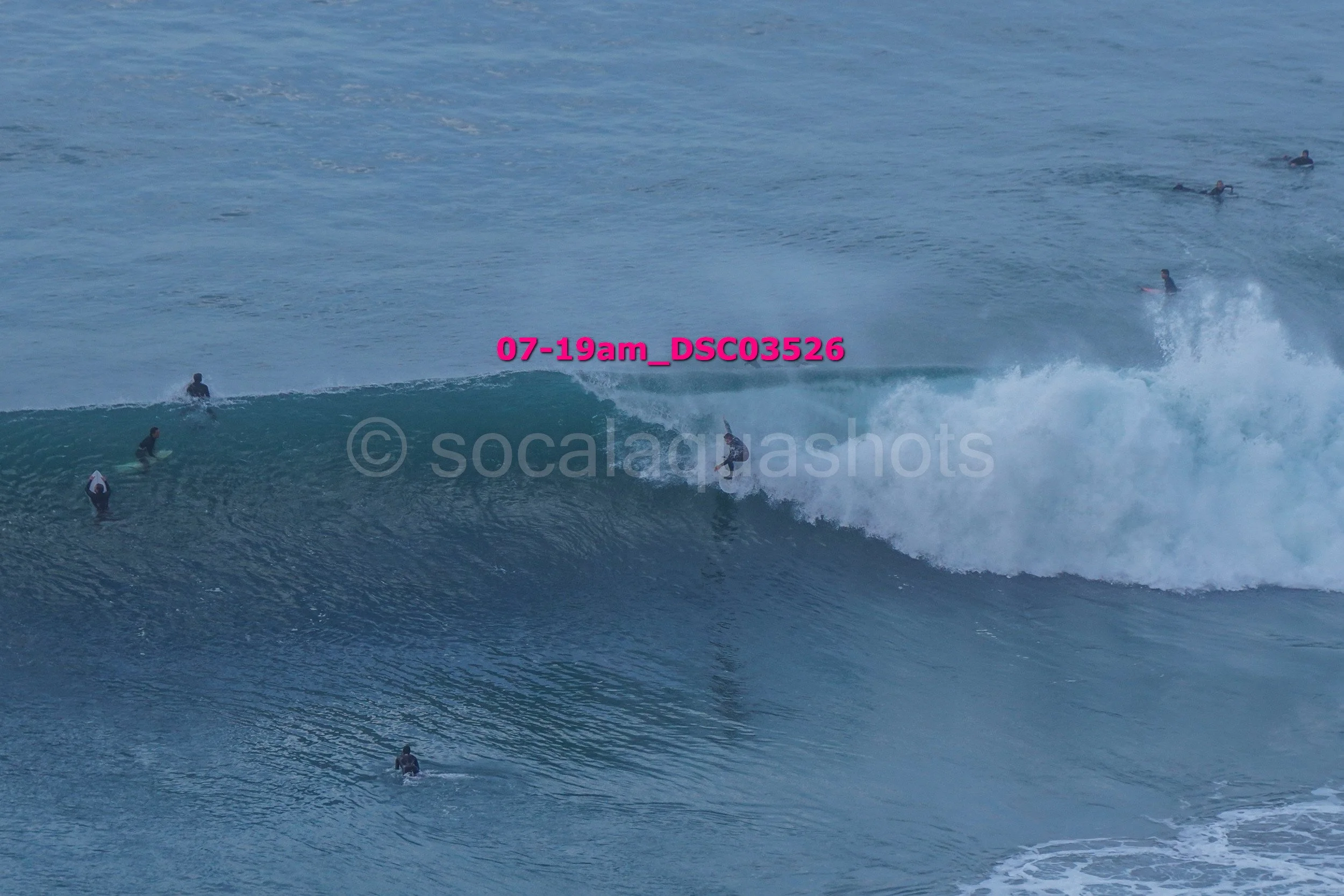 A group of surfers in the ocean, with some riding a large wave and others waiting in the water for their turn, under a clear blue sky.
