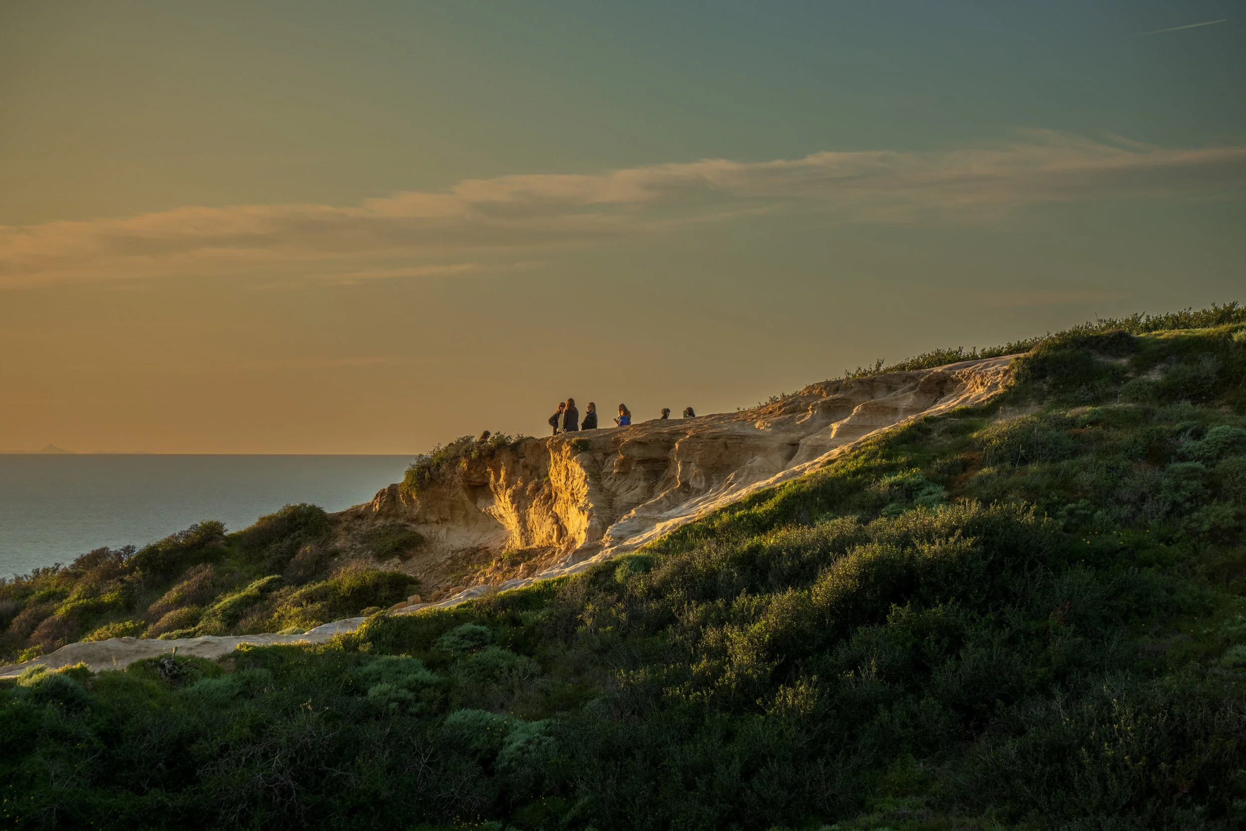 Group of people standing on a rocky hill overlooking the ocean at sunset.
