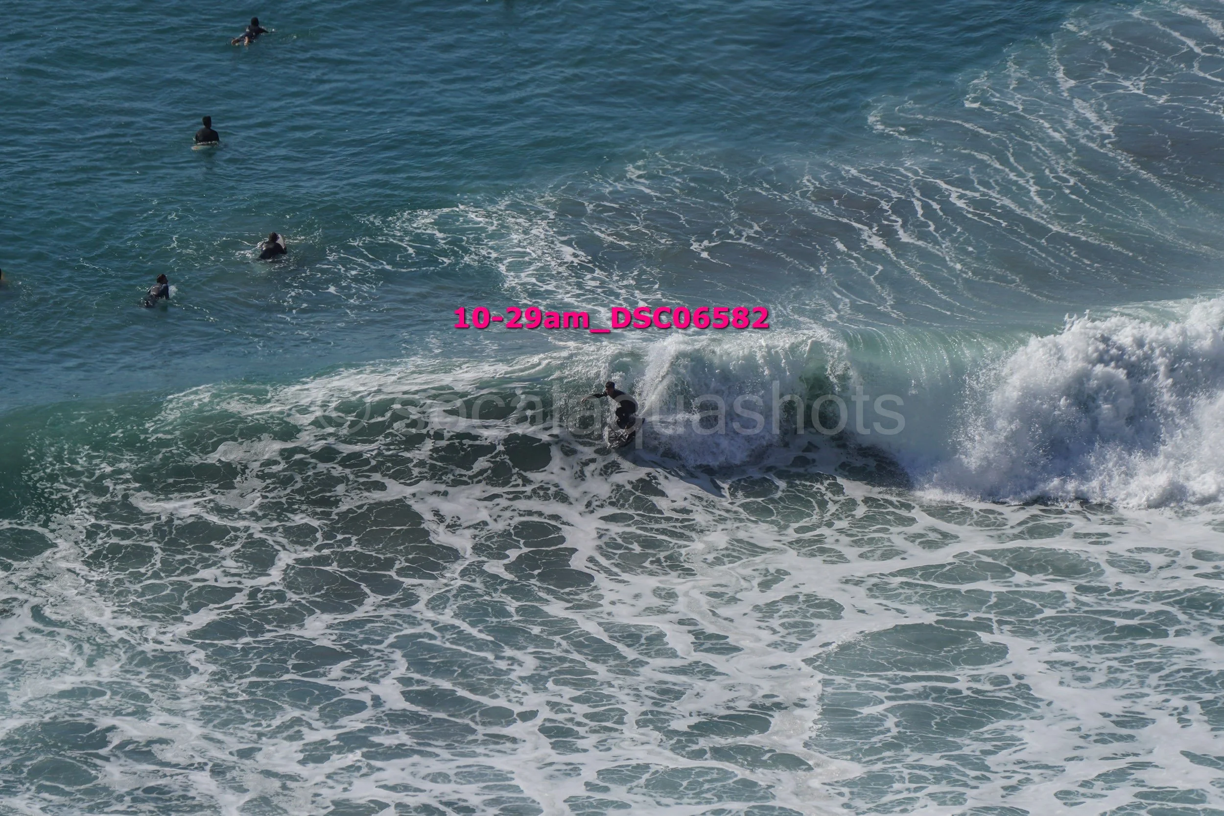 People surfing in the ocean with waves breaking, several individuals swimming or waiting in the water.