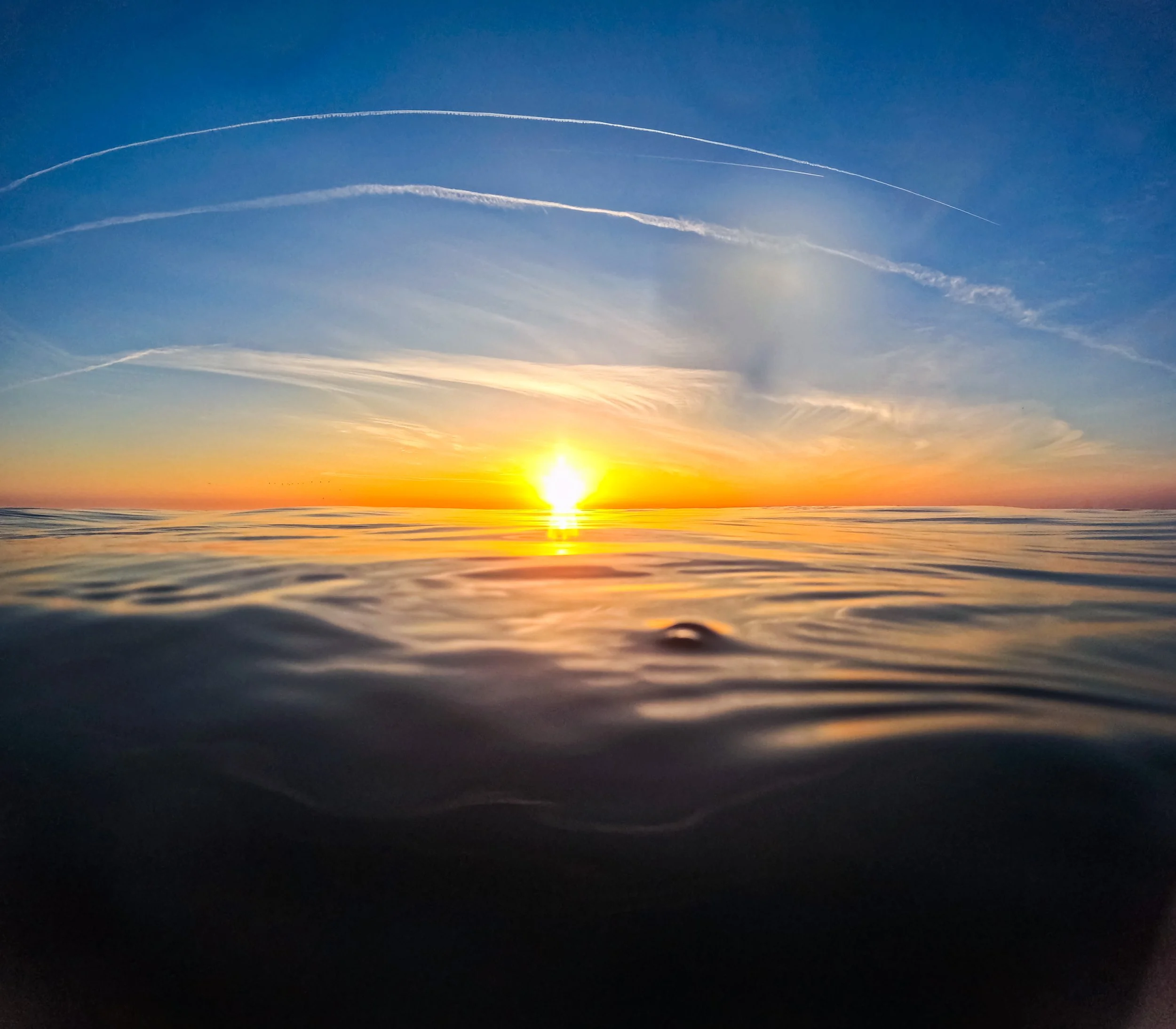 Sunset over calm ocean waters with colorful sky and contrails.