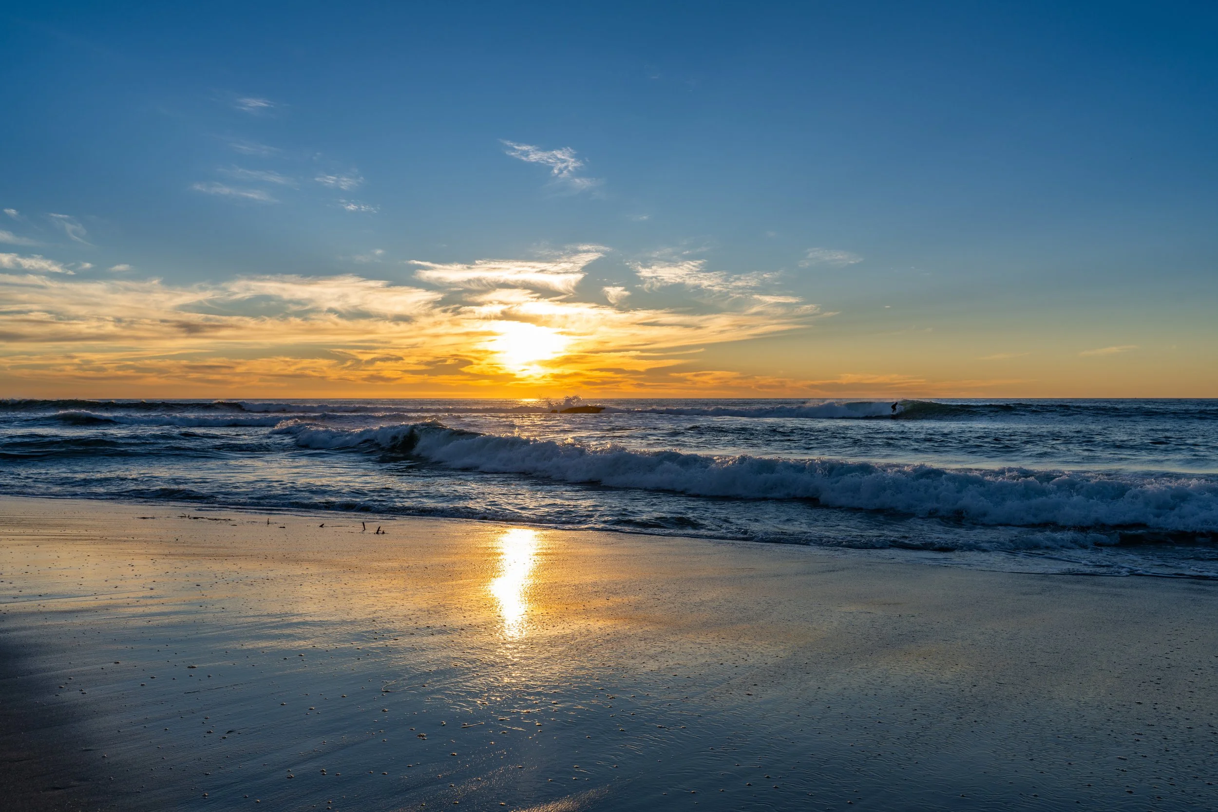 Sunset over the ocean with waves crashing on the sandy beach and a bright reflection of the sun on the water.