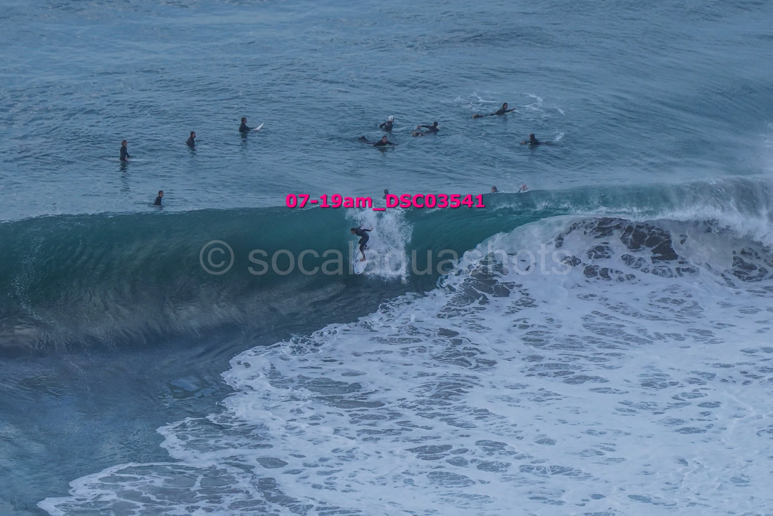 A person surfing on a large wave near the shoreline with several people swimming and floating in the water in the background.
