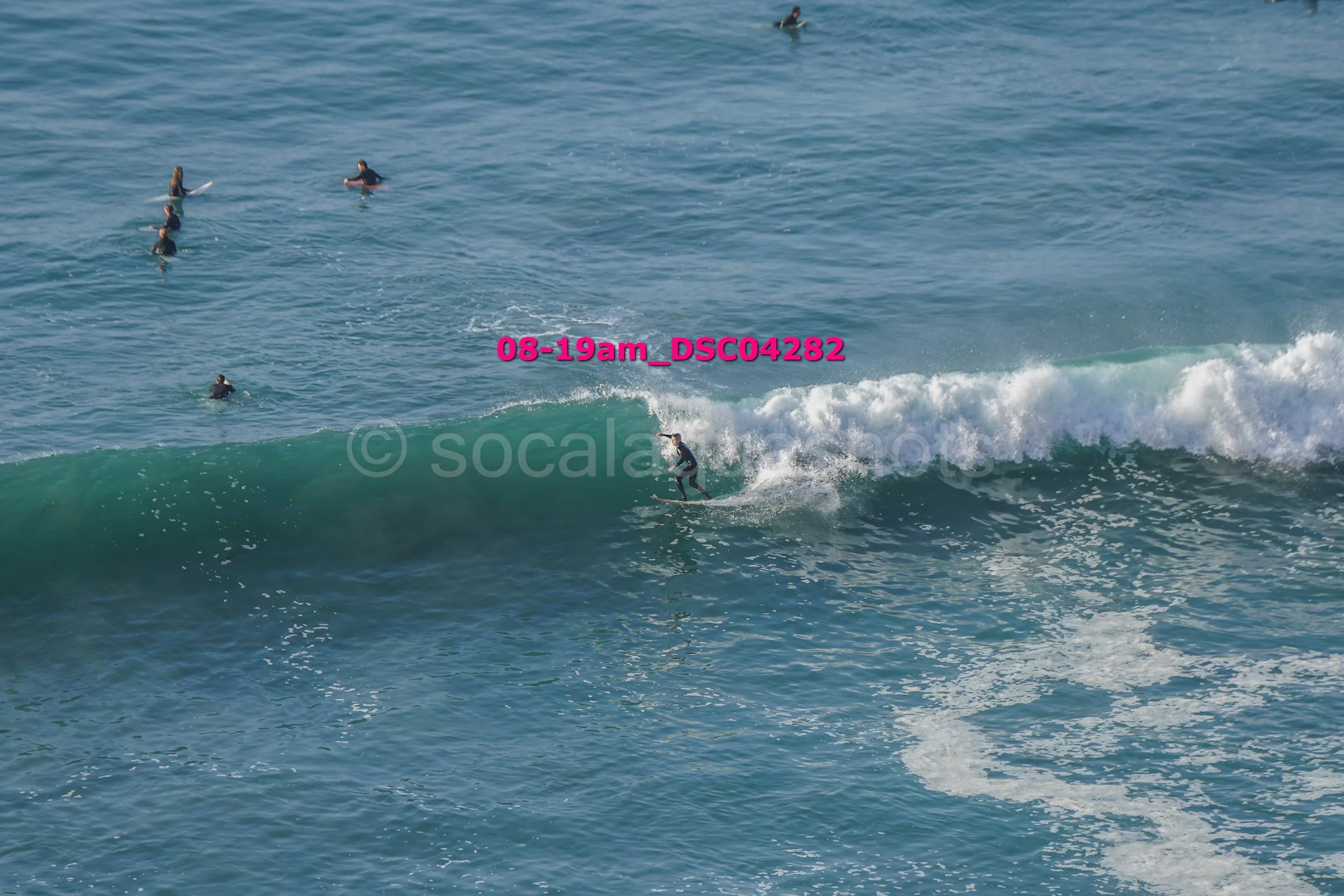 Person surfing on a wave in the ocean with several people swimming in the background.