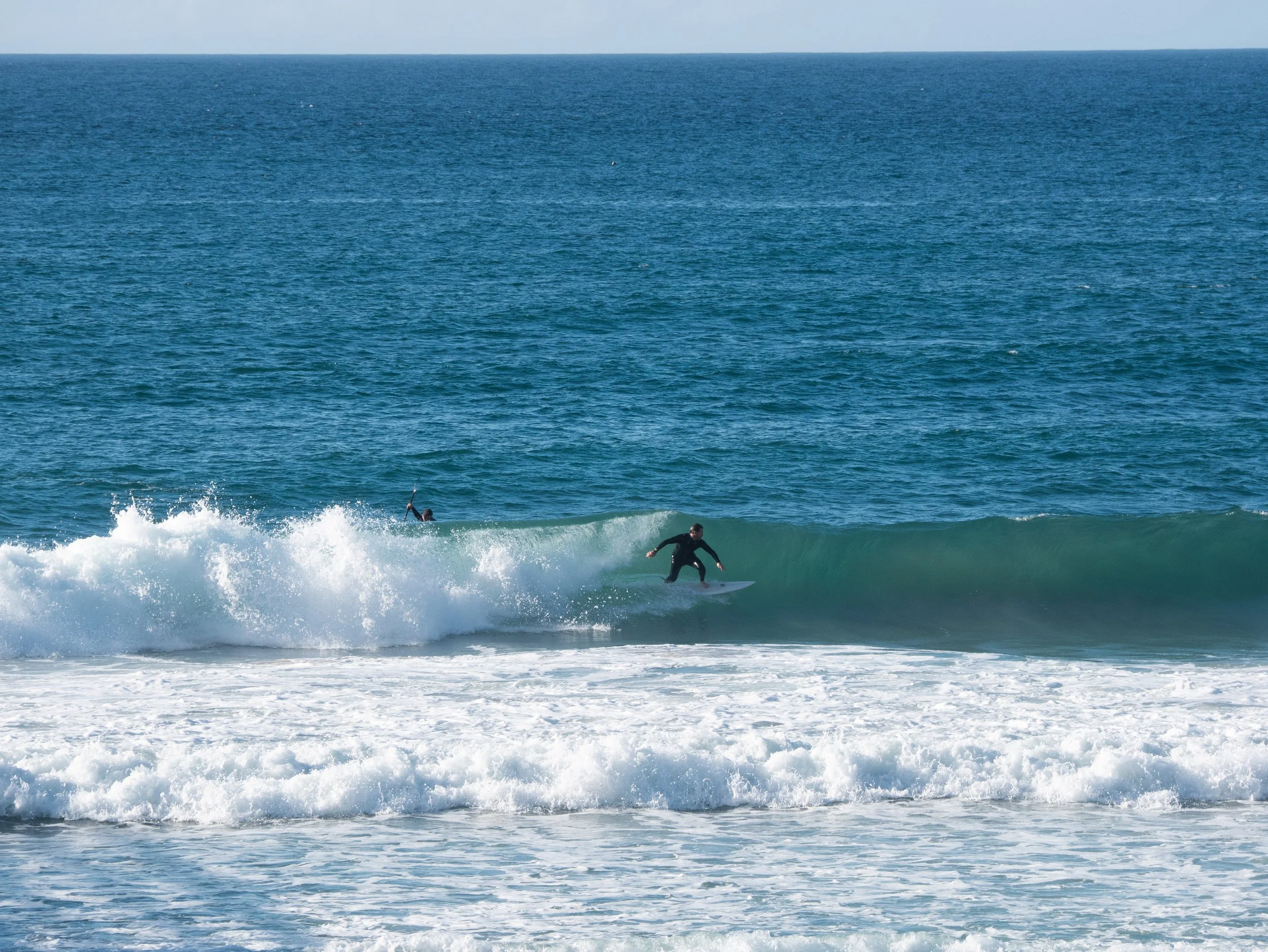 A person surfing on a small wave in the ocean with another person swimming in the background.