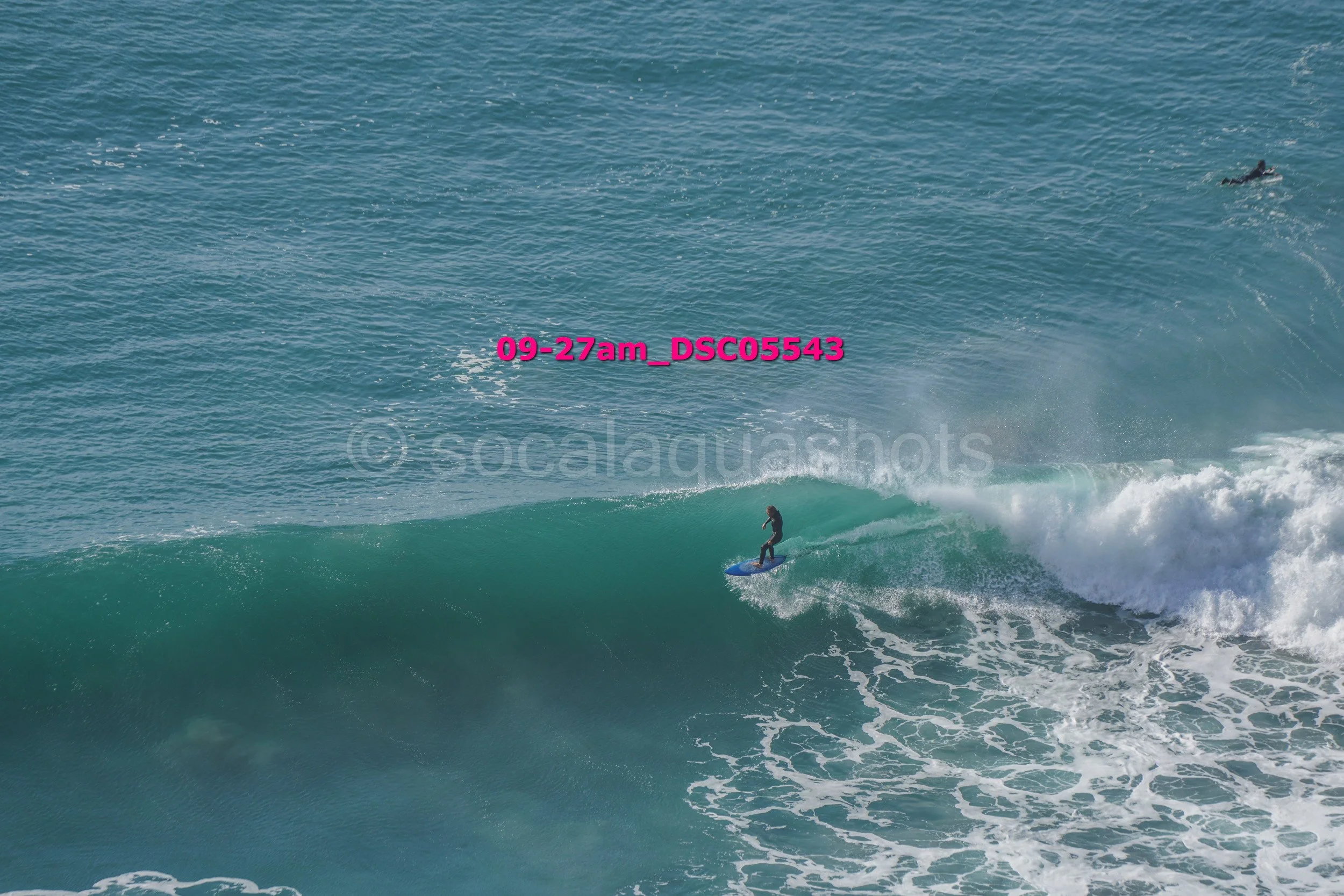 A surfer riding a wave in the ocean with another person swimming in the water nearby, bright blue sky.