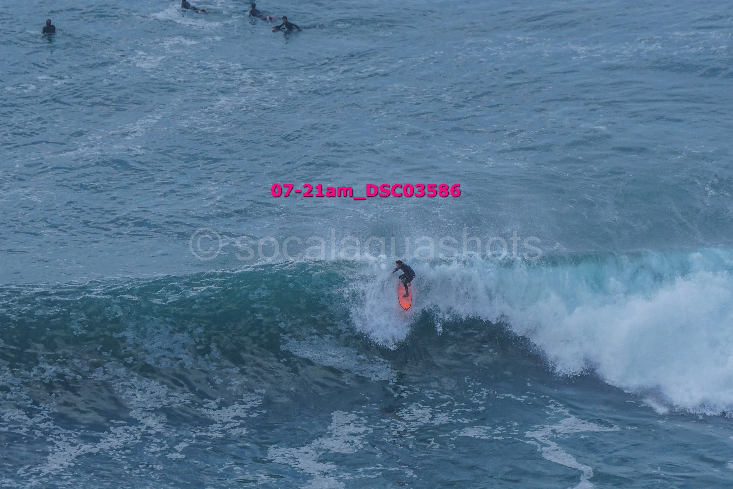 A person surfing on a large wave in the ocean, with several other surfers visible in the background.