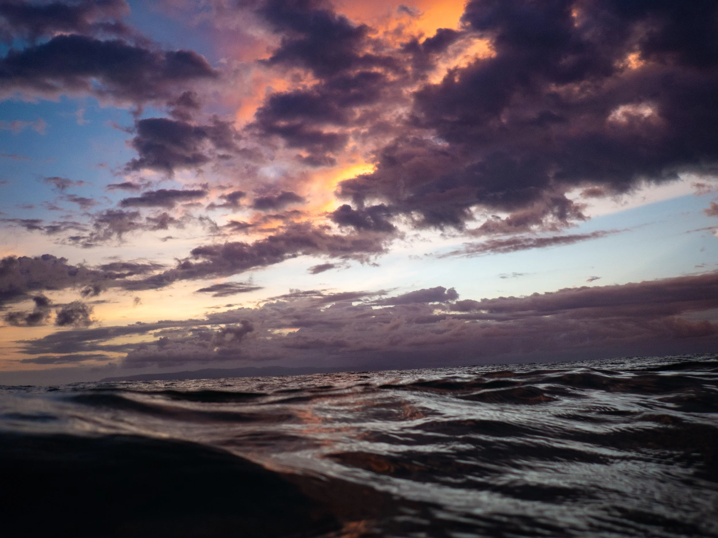 A scenic view of a cloudy sky during sunset over the ocean, with the water reflecting the colors of the sky and waves in the foreground.