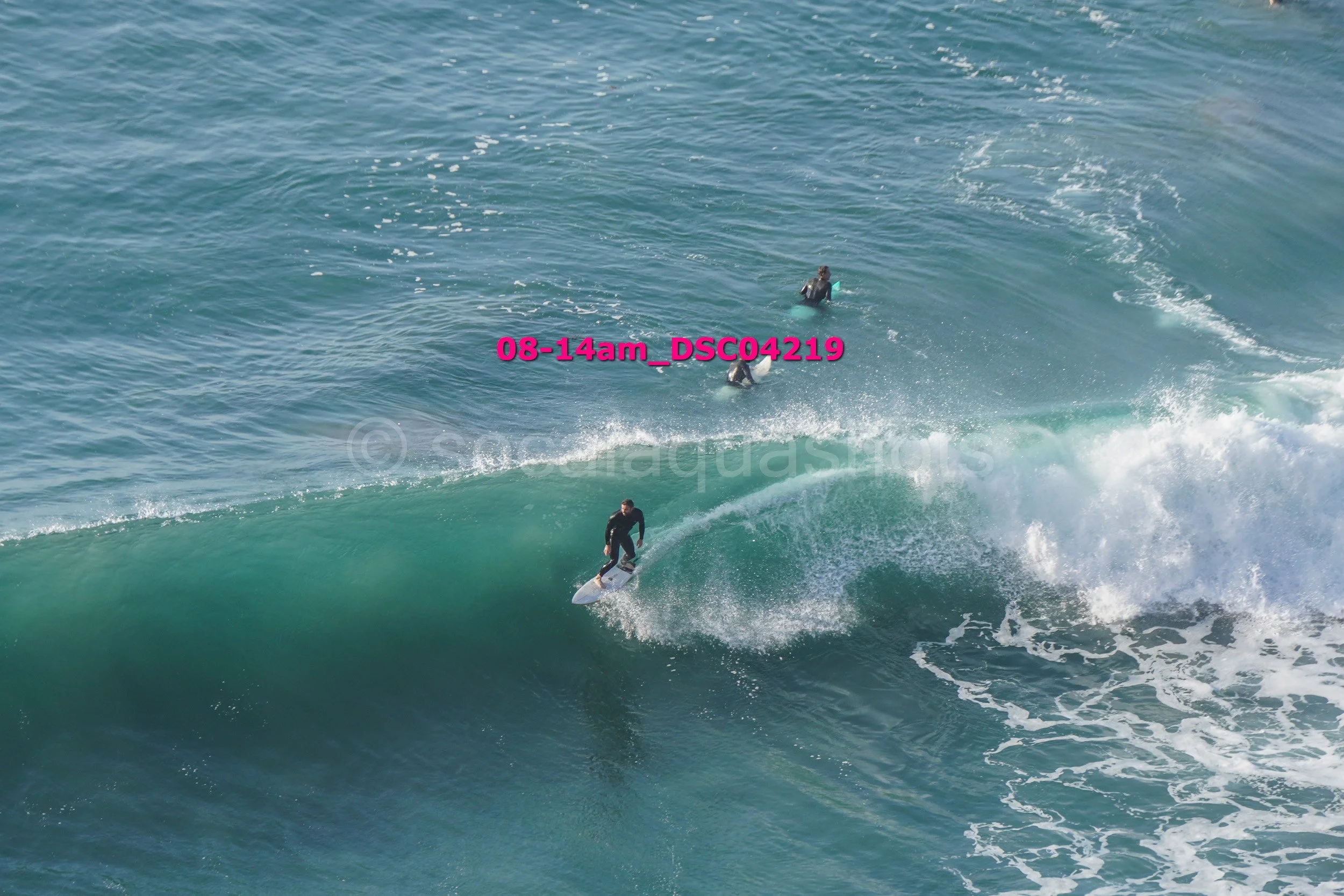 A person surfing on a large wave with two other surfers visible in the background in the ocean.