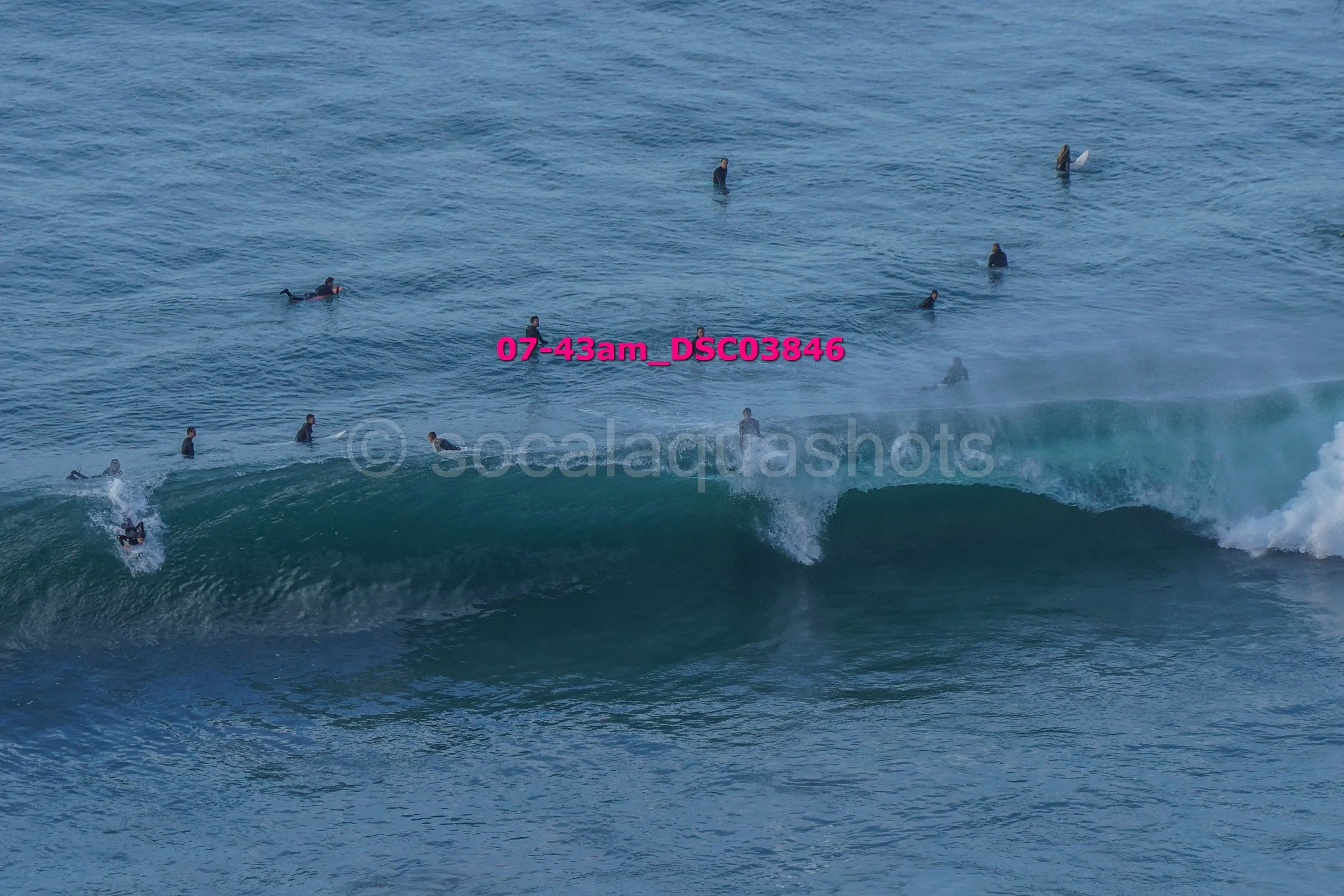 A group of surfers in wetsuits, some on surfboards and others in the water, waiting for or riding a wave in the ocean during daytime.
