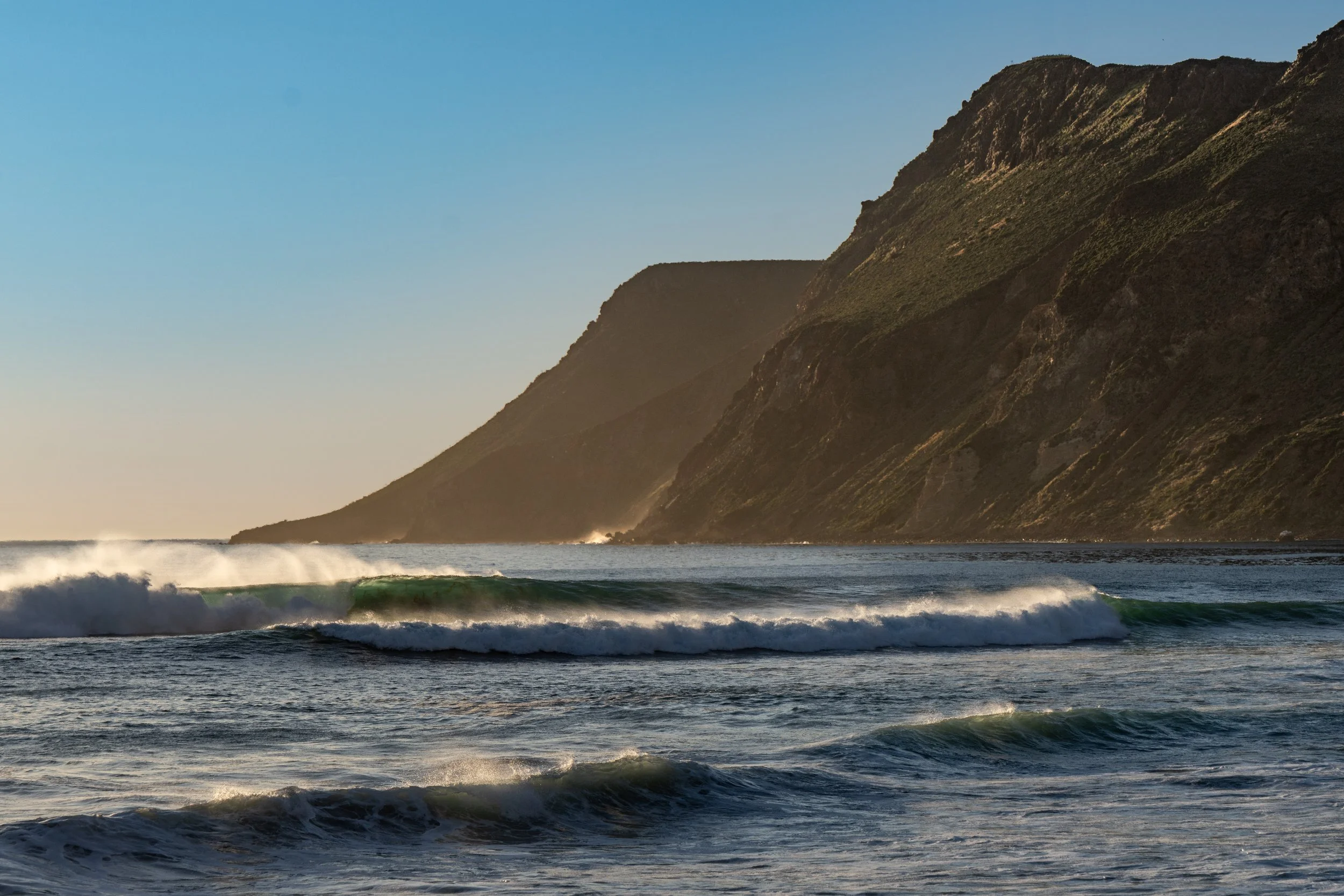 Ocean waves crashing near a coastline with steep cliffs and green vegetation under a clear blue sky.