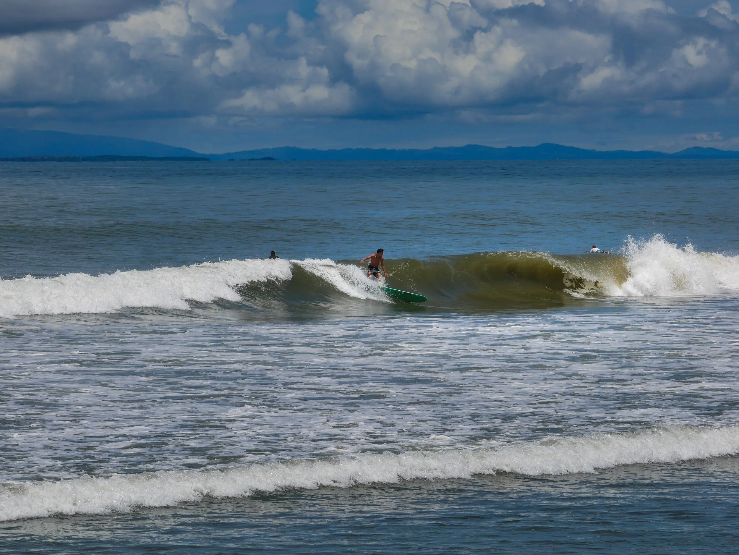 Surfer riding a wave in the ocean with cloudy sky and distant hills in the background.