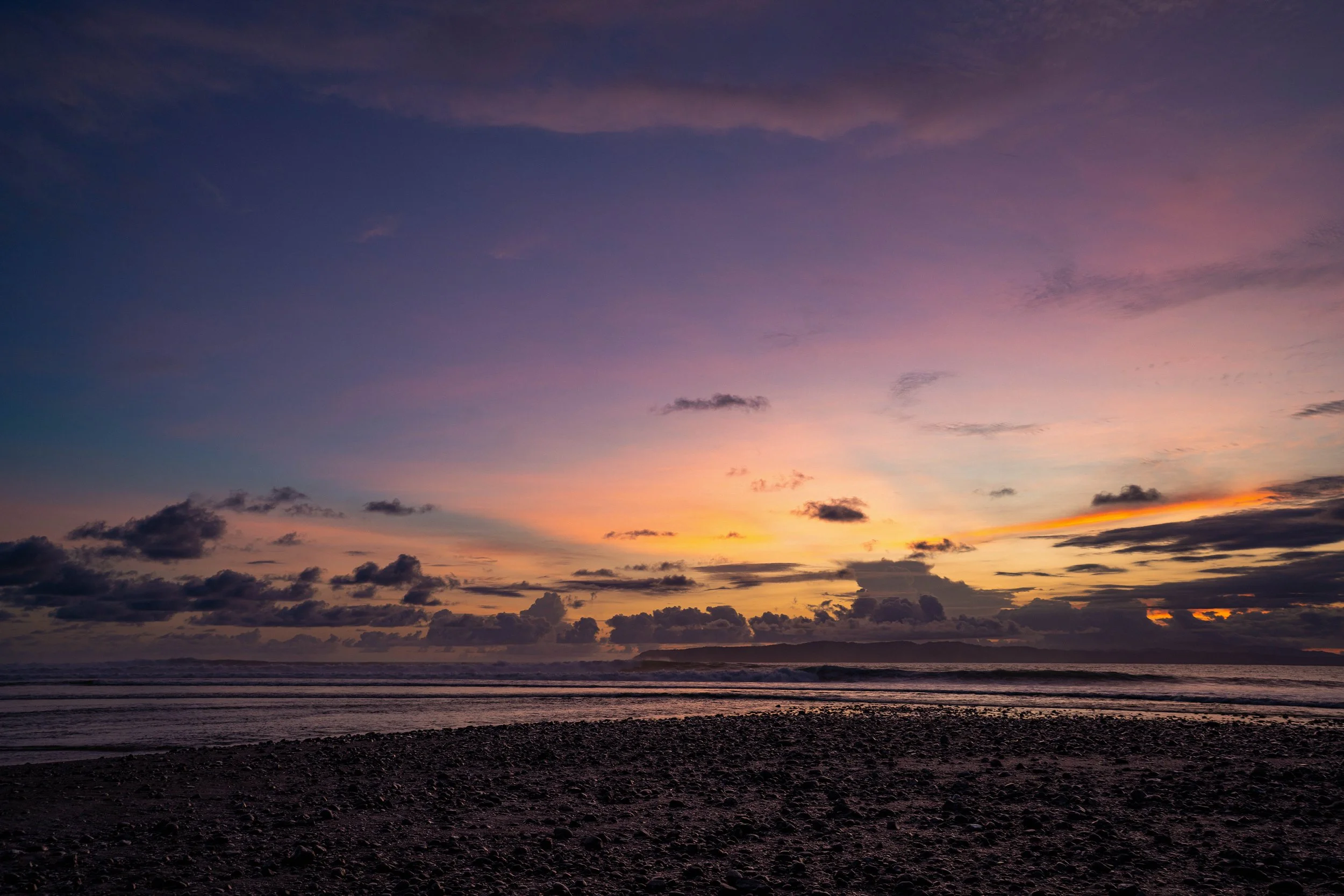 Sunset over the ocean with purple, pink, and orange hues in the sky, dark clouds near the horizon, and a rocky shoreline in the foreground.