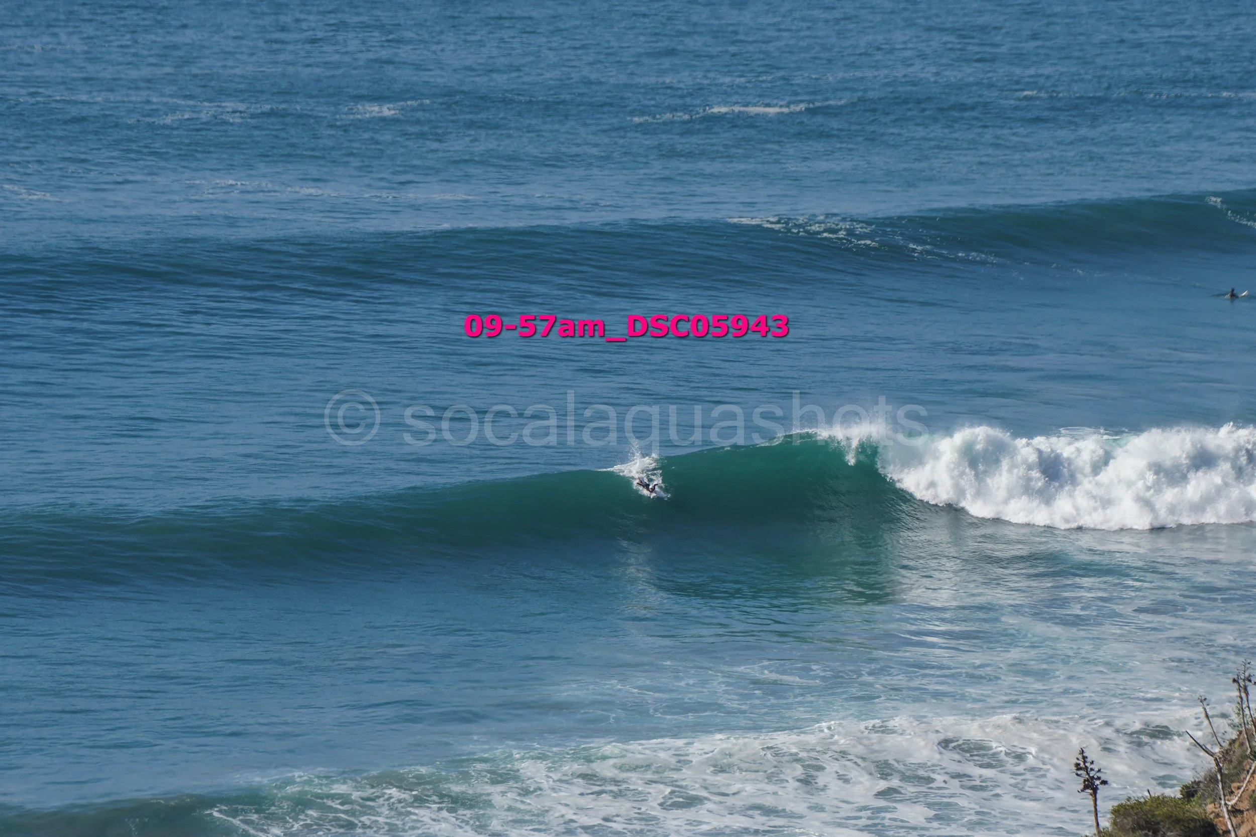 A surfer riding a small wave in the ocean near the shore, with a few other surfers visible further out. The ocean has gentle waves and the sky is clear.