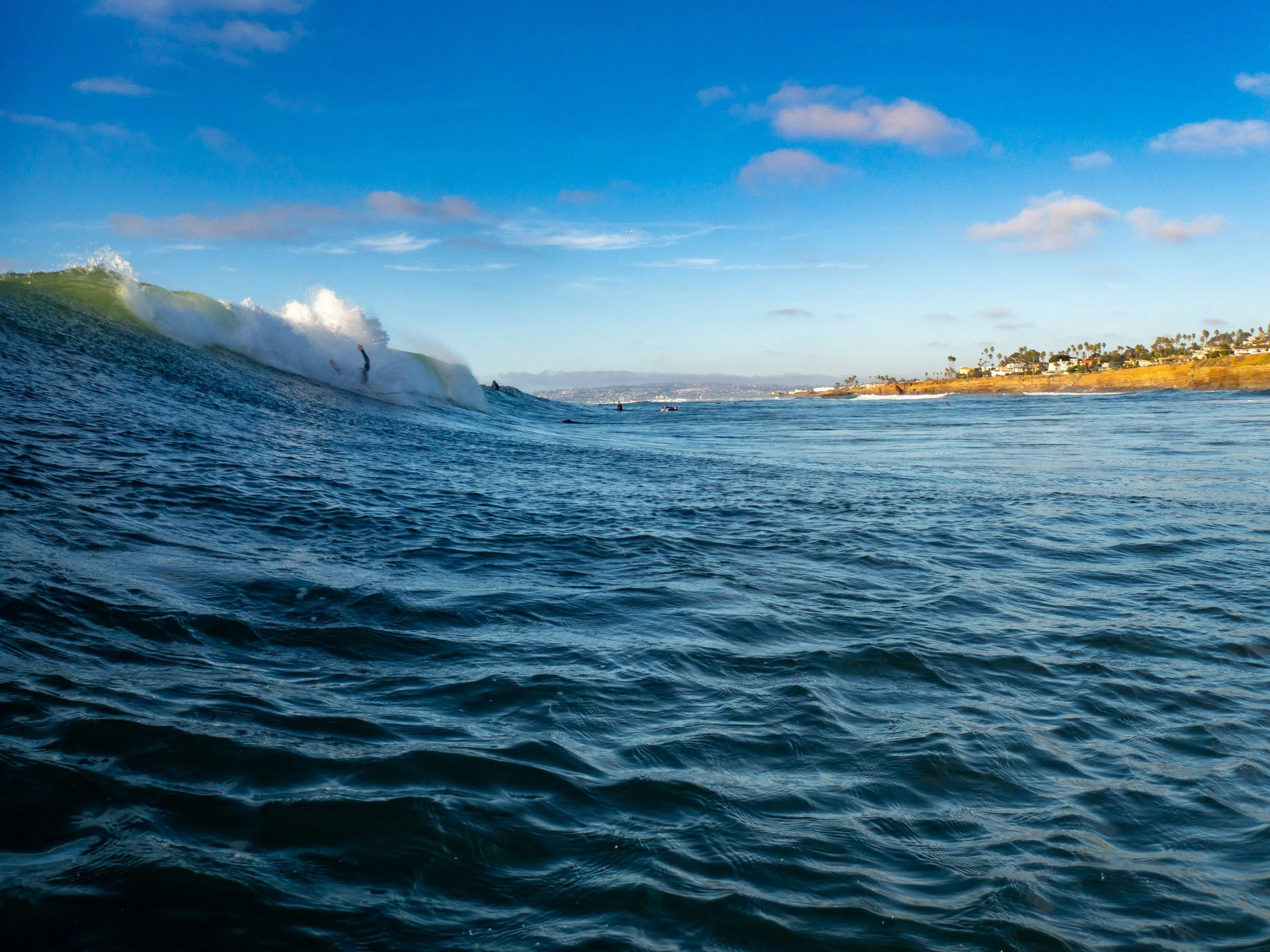Ocean view with a large wave and surfers, coastline with houses and palm trees under a partly cloudy sky.