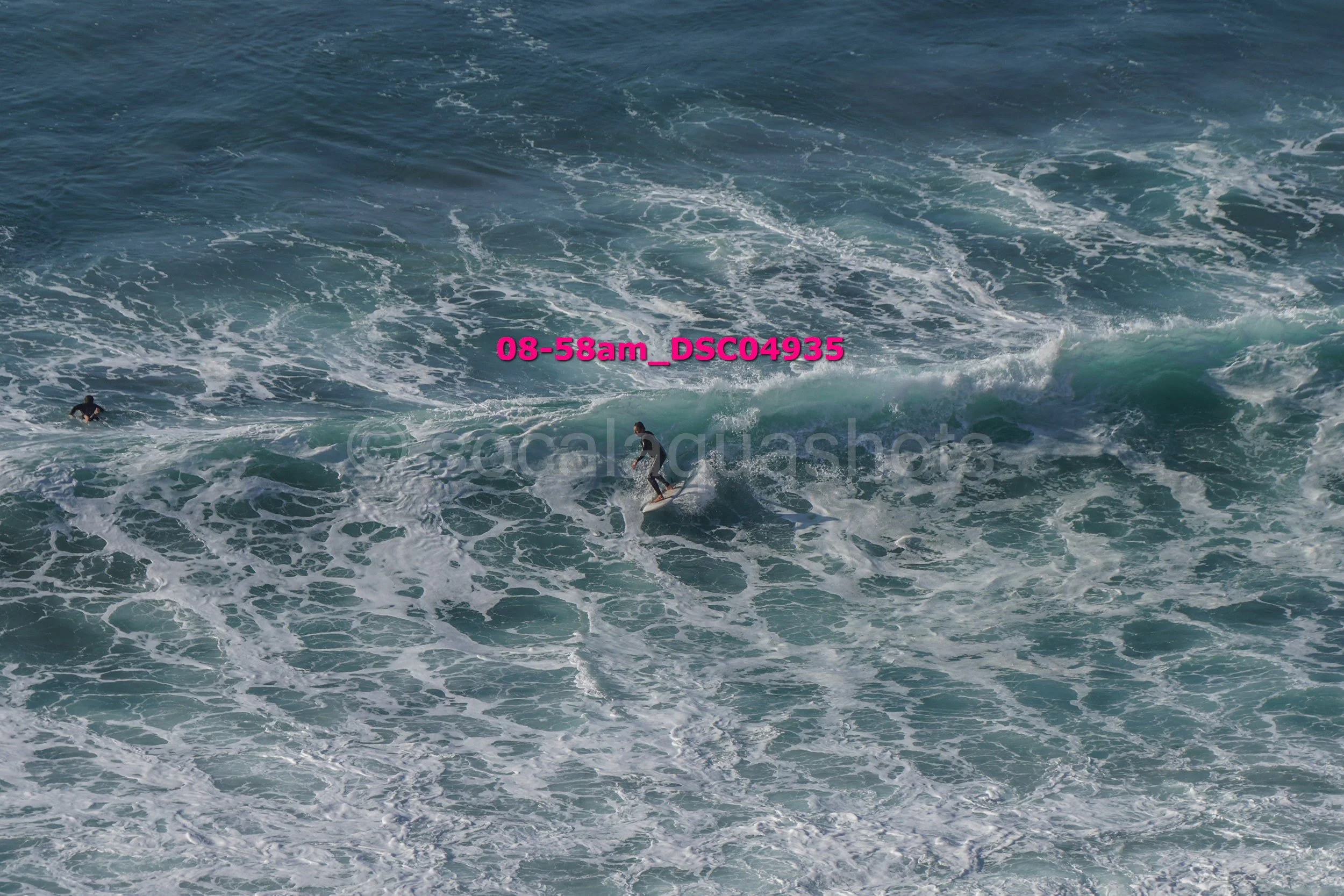 Surfer riding a wave in the ocean with another surfer swimming nearby.