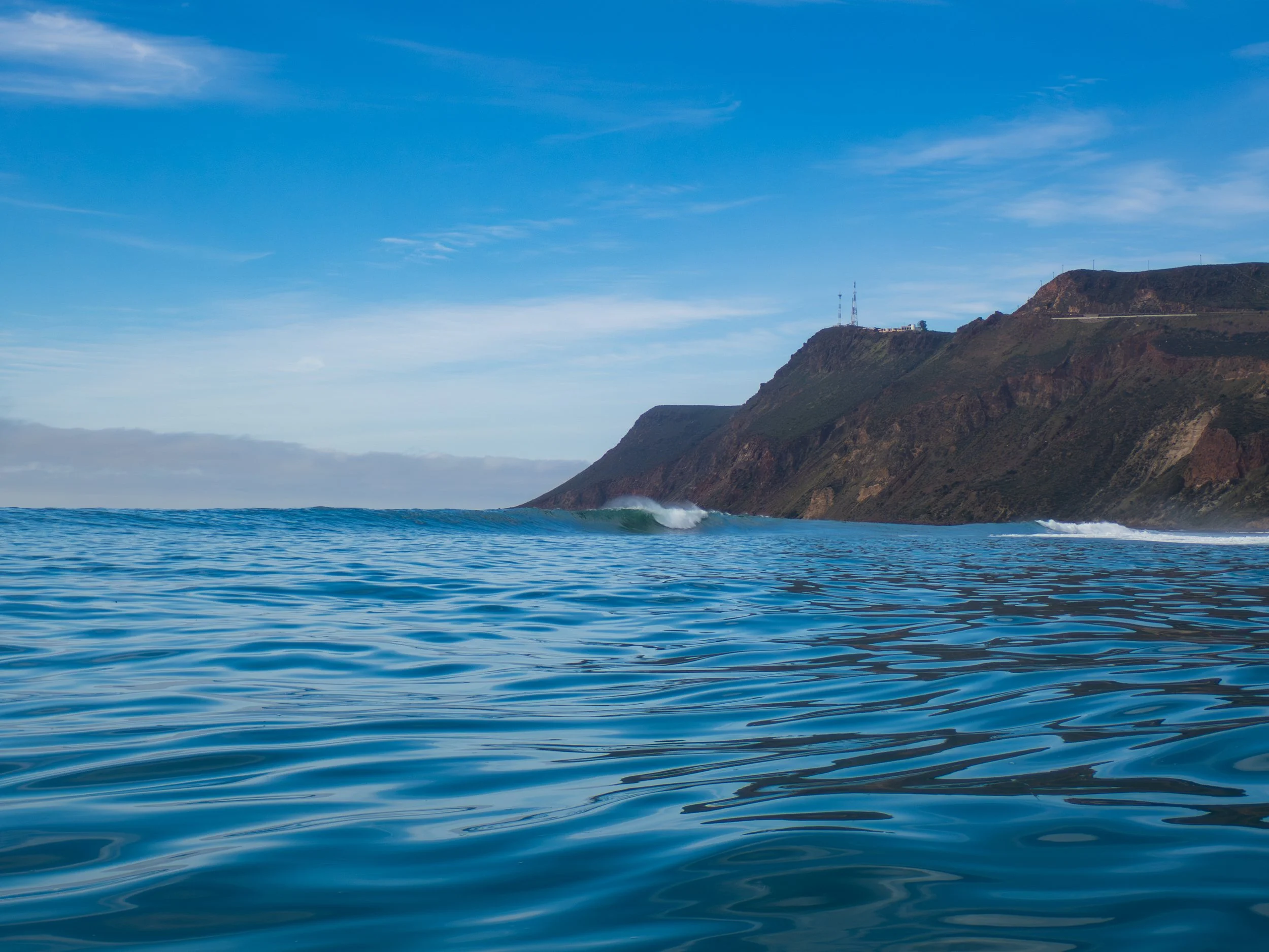 Calm ocean water with small waves, rocky cliffside in the background, and a partly cloudy sky.