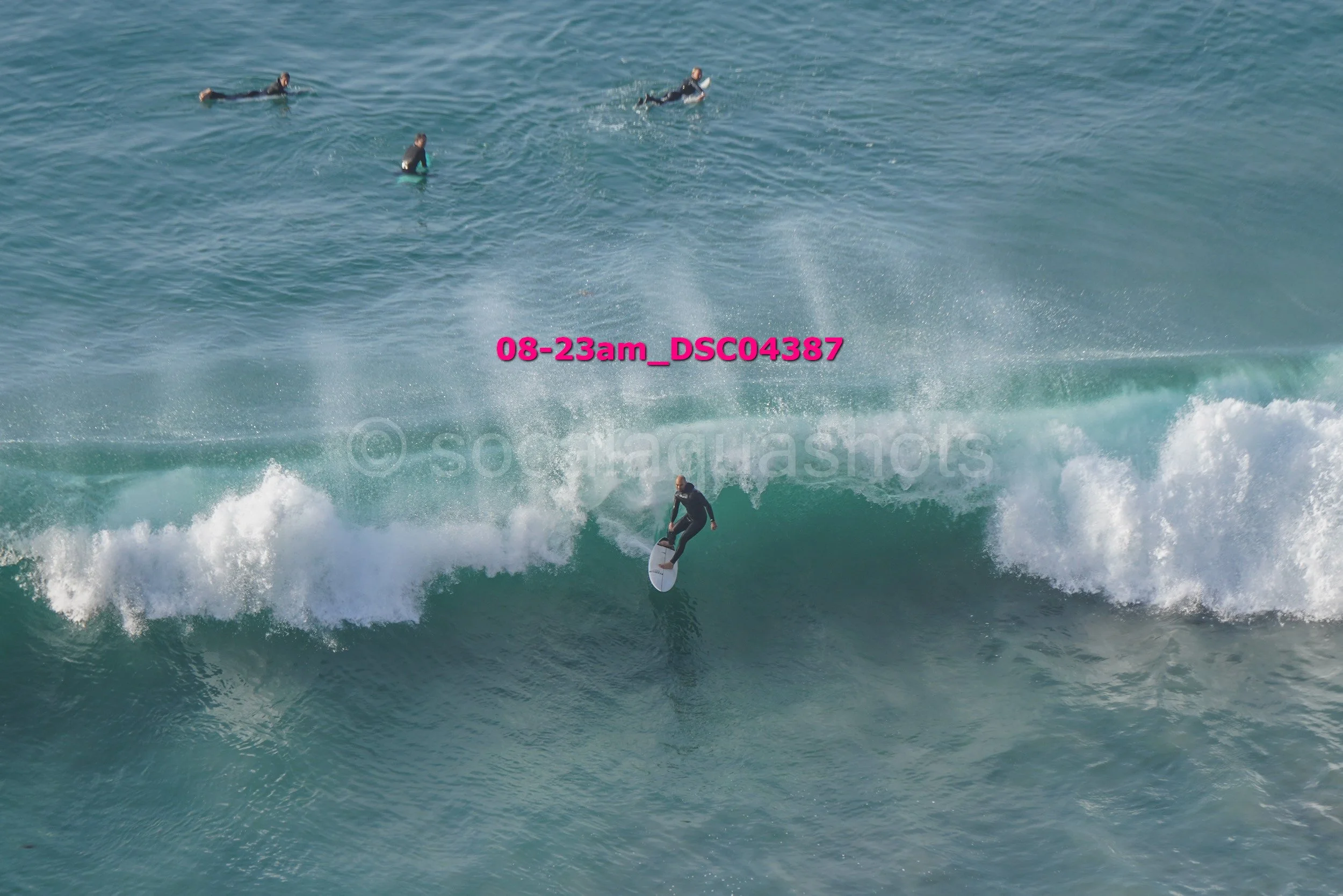 A person surfing on a wave with three other people swimming or floating in the water nearby.