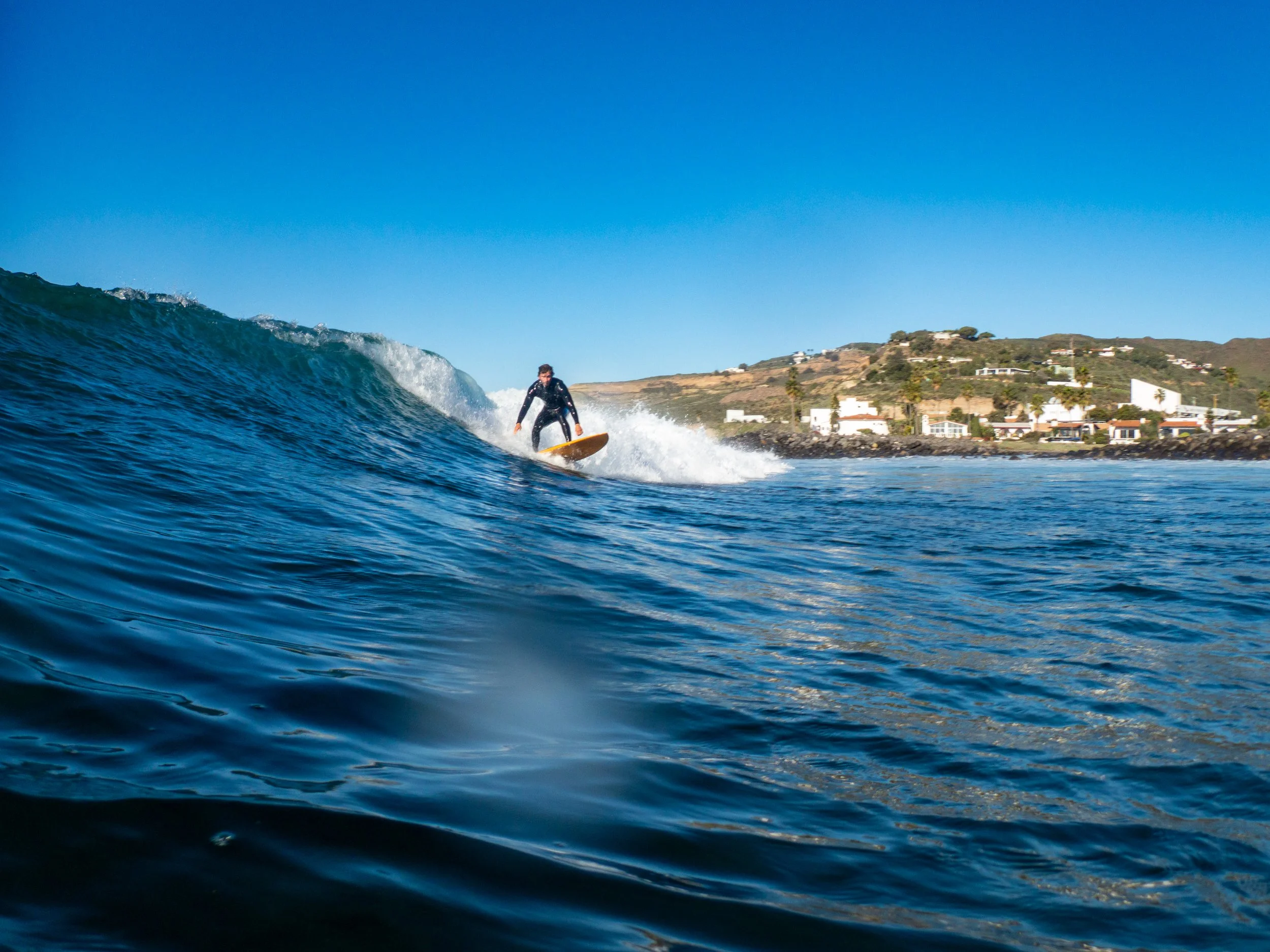 A person surfing on a blue wave near the coast, with houses and hills visible in the background under a clear blue sky.