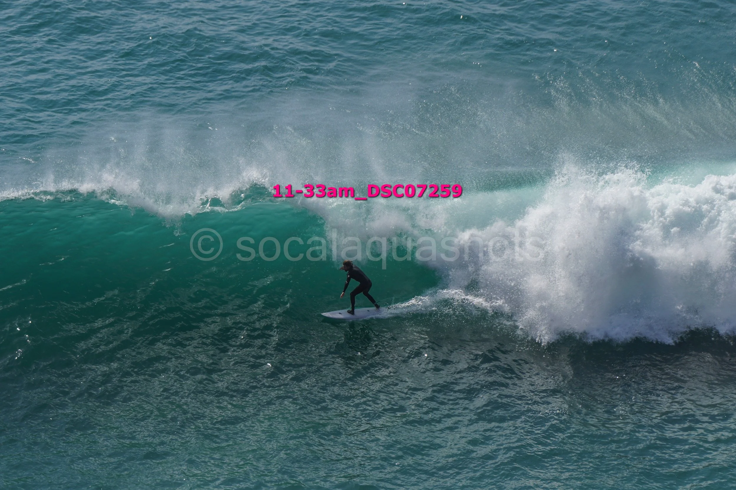 A person surfing on a large ocean wave, wearing a black wetsuit, with water spray and foam around.