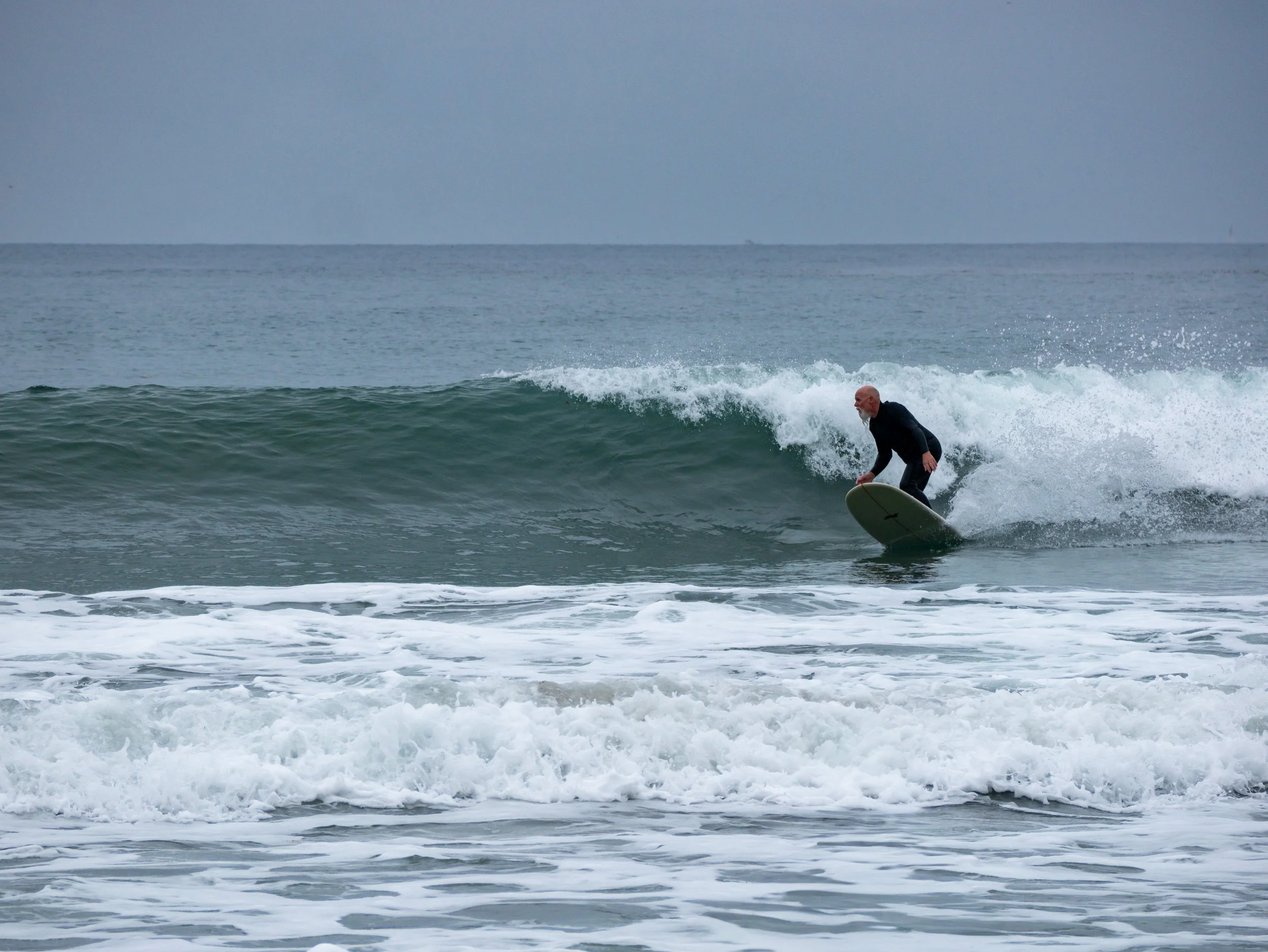 A man surfing on a small wave in the ocean, wearing a black wetsuit, with a cloudy sky in the background.