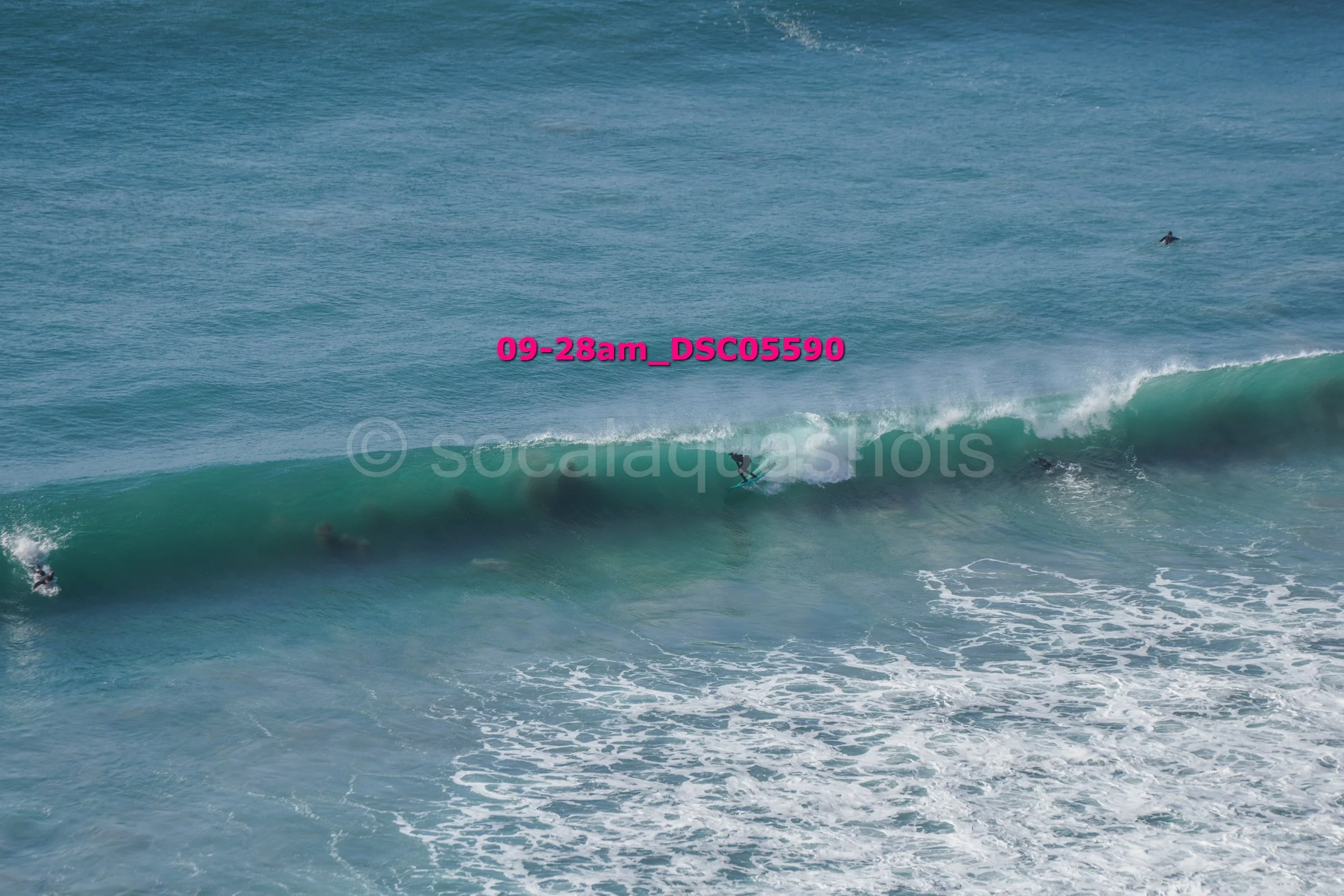 Surfer riding a wave in the ocean with two other surfers paddling nearby and one further away, under a clear sky.