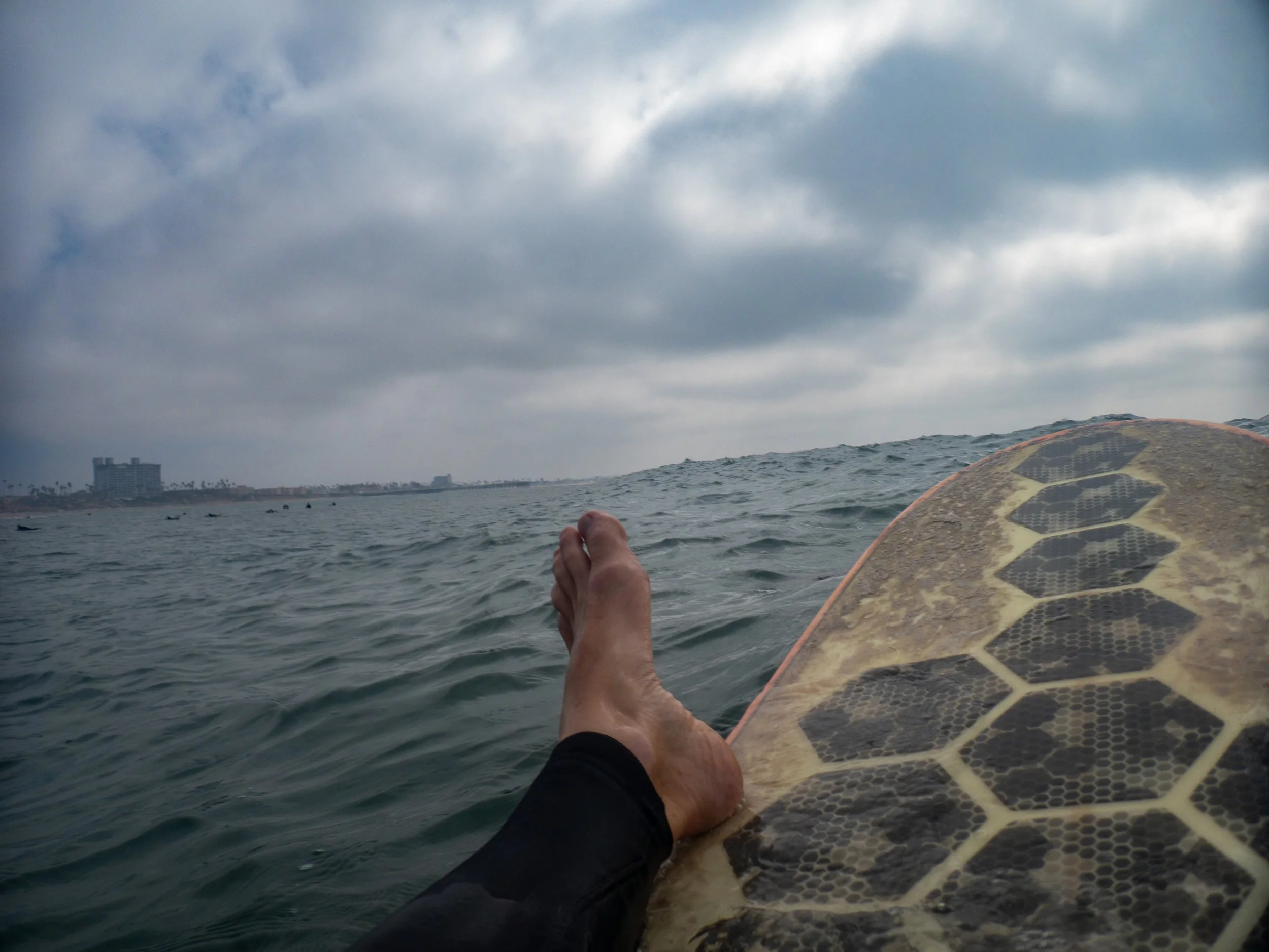 A person cooling off while lying on a surfboard in the ocean, with a cloudy sky above and city buildings in the distance.