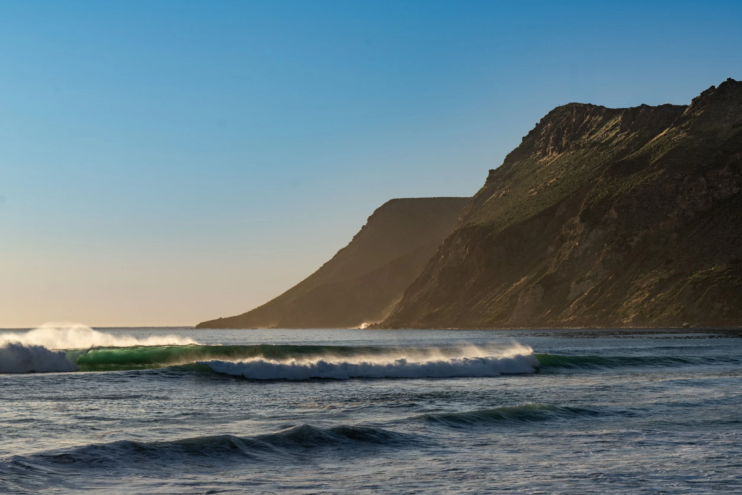 Ocean waves crashing on the shore with mountainous cliffs in the background during sunset.