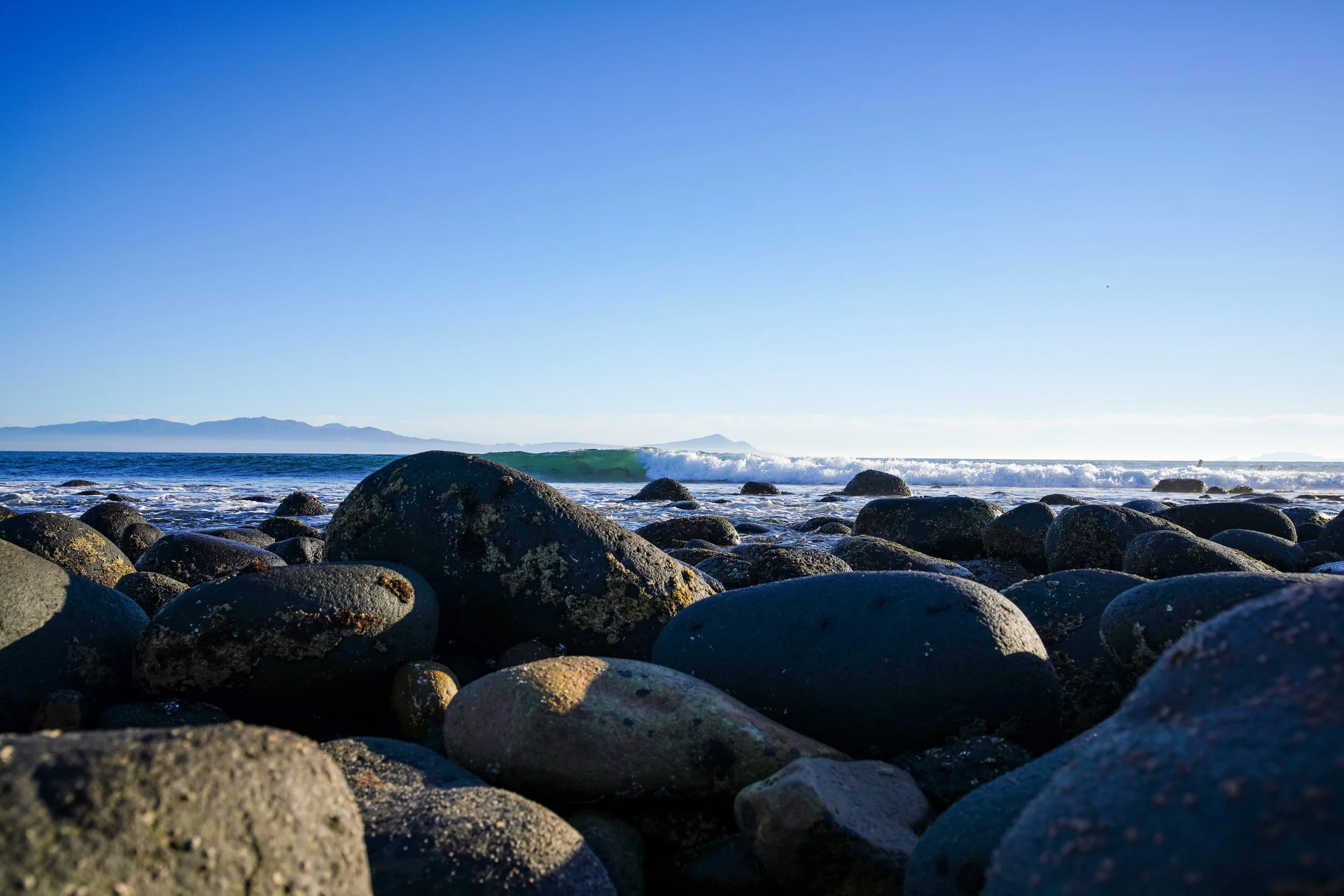 A rocky beach with large dark stones in the foreground, ocean waves, and distant islands under a clear blue sky.