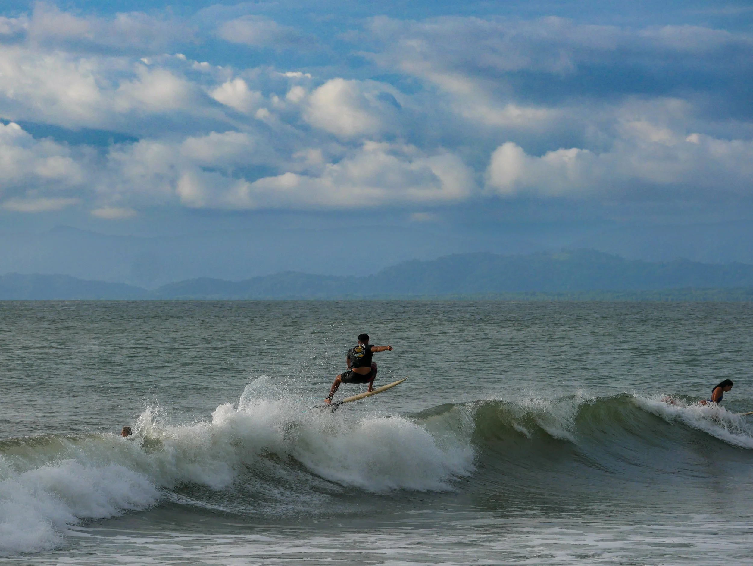Surfer riding a wave in the ocean with cloudy sky and mountains in the background.