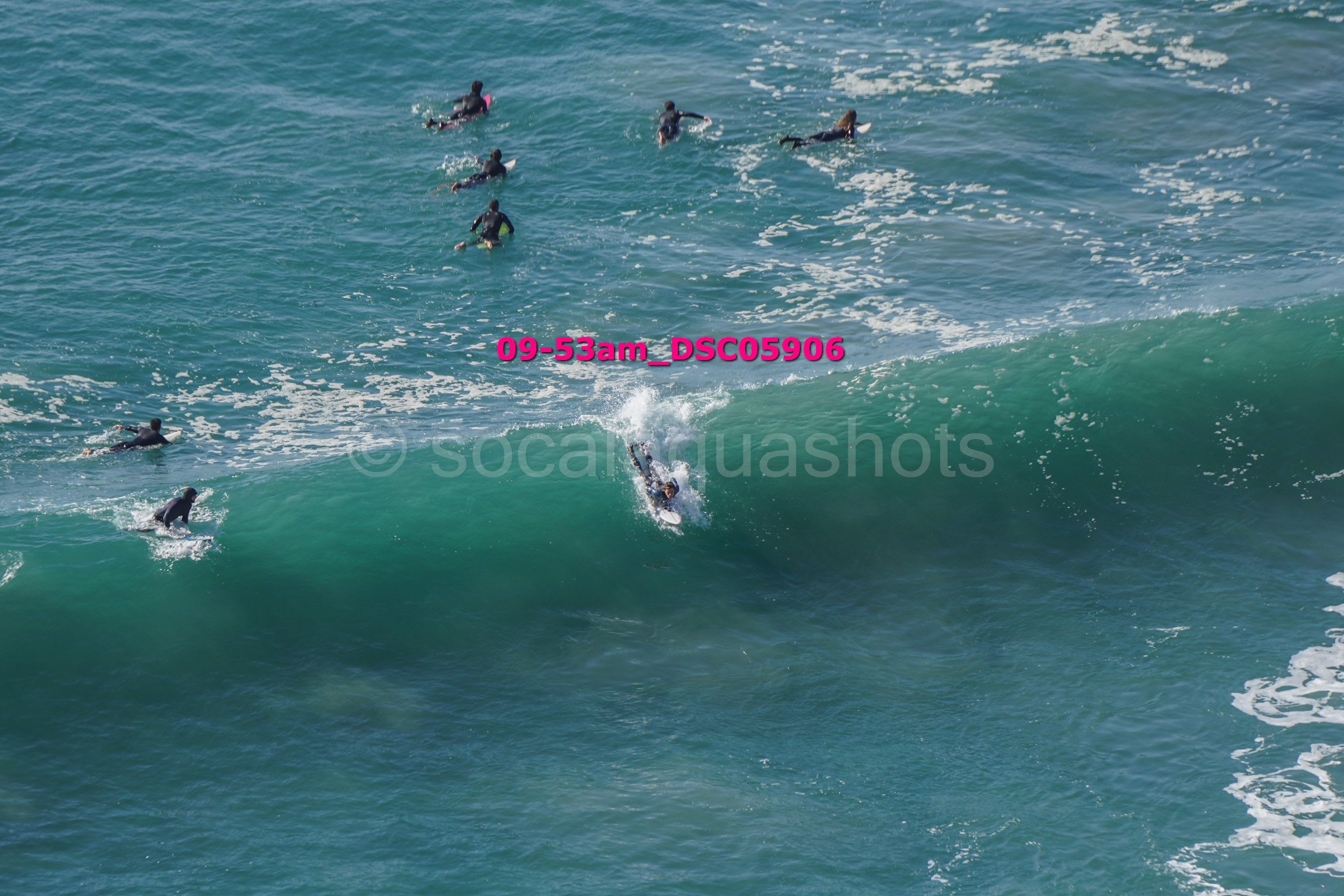 Group of surfers riding a large wave in the ocean, some paddling and others standing on surfboards.