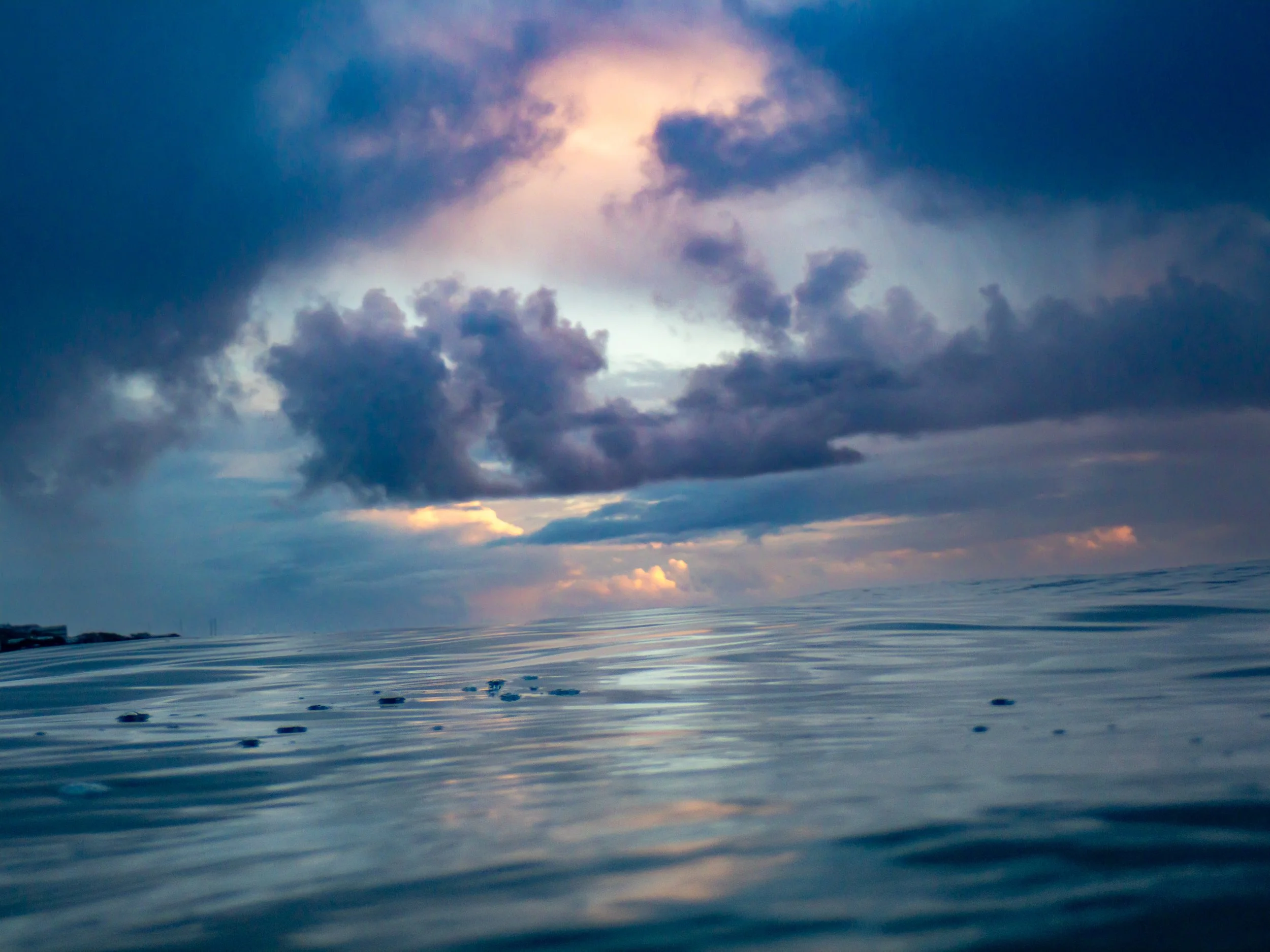 View of calm ocean water reflecting a cloudy sky with pink and orange hues at sunset or sunrise.