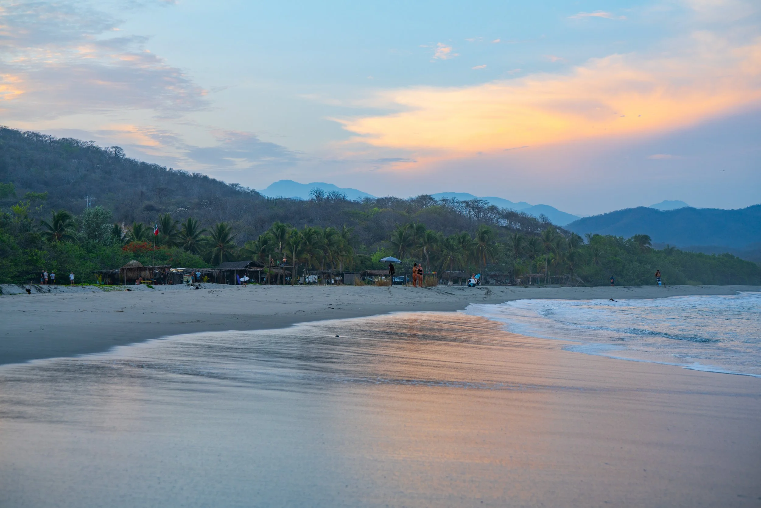 Sunset over a beach with calm waves, palm trees, and a small group of people near the shore, mountains in the background.