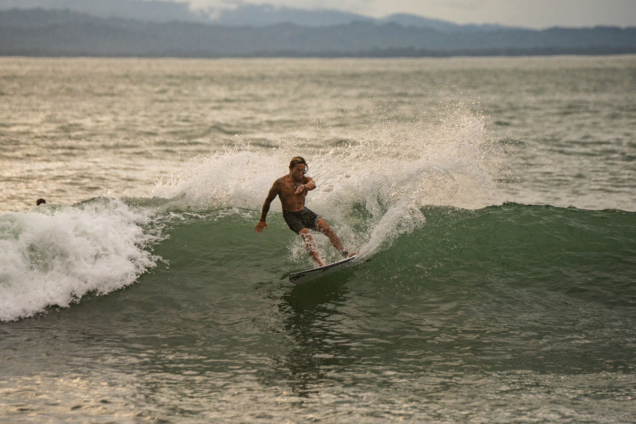 A person surfing on a wave in the ocean, with mountains visible in the distant background.