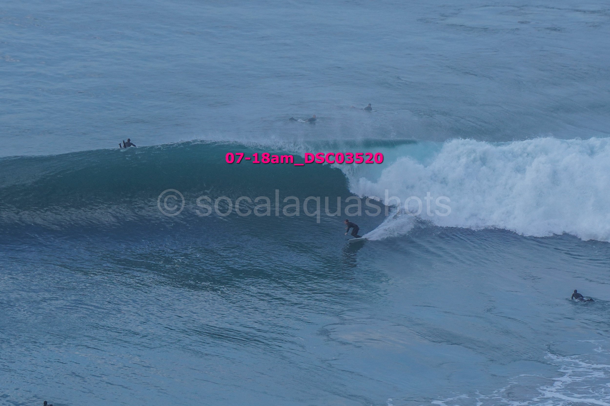 Surfer riding a large ocean wave with several other surfers in the water nearby.